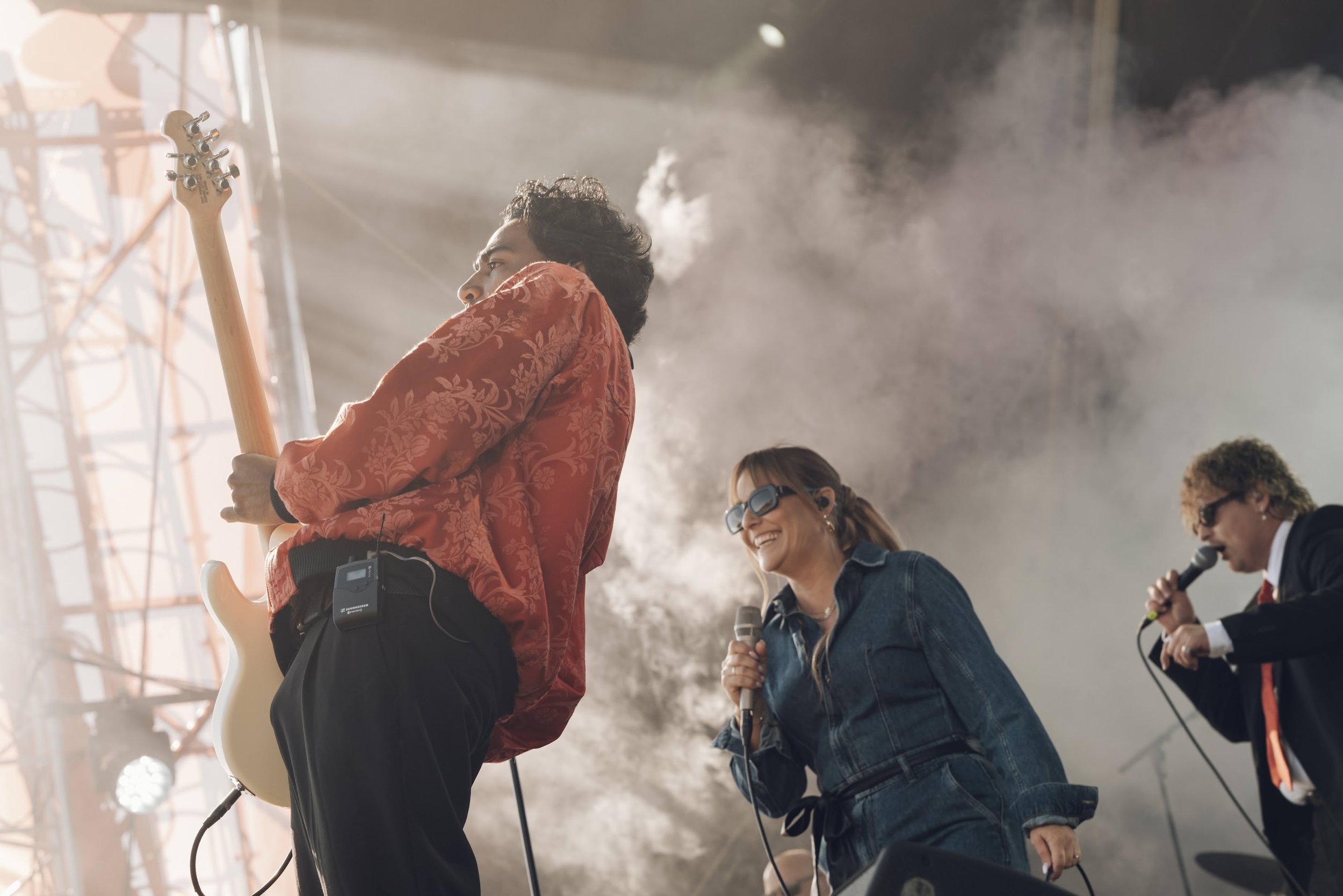 Tre musikere på scenen under en konsert med lysspotlights og røyk, en mann spiller gitar, en kvinne synger med mikrofon og en mann synger med mikrofon, alle smilende.