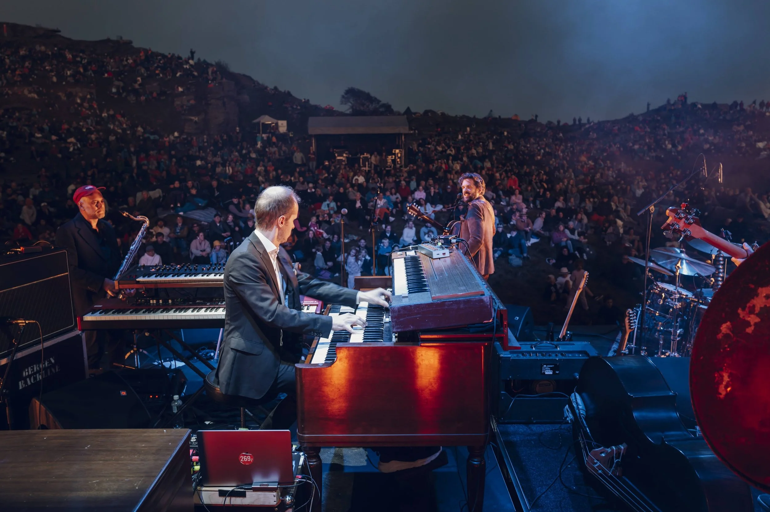 Konsert med fire musikere på scen med et stort publikum i bakgrunnen, sett fra bakkant.