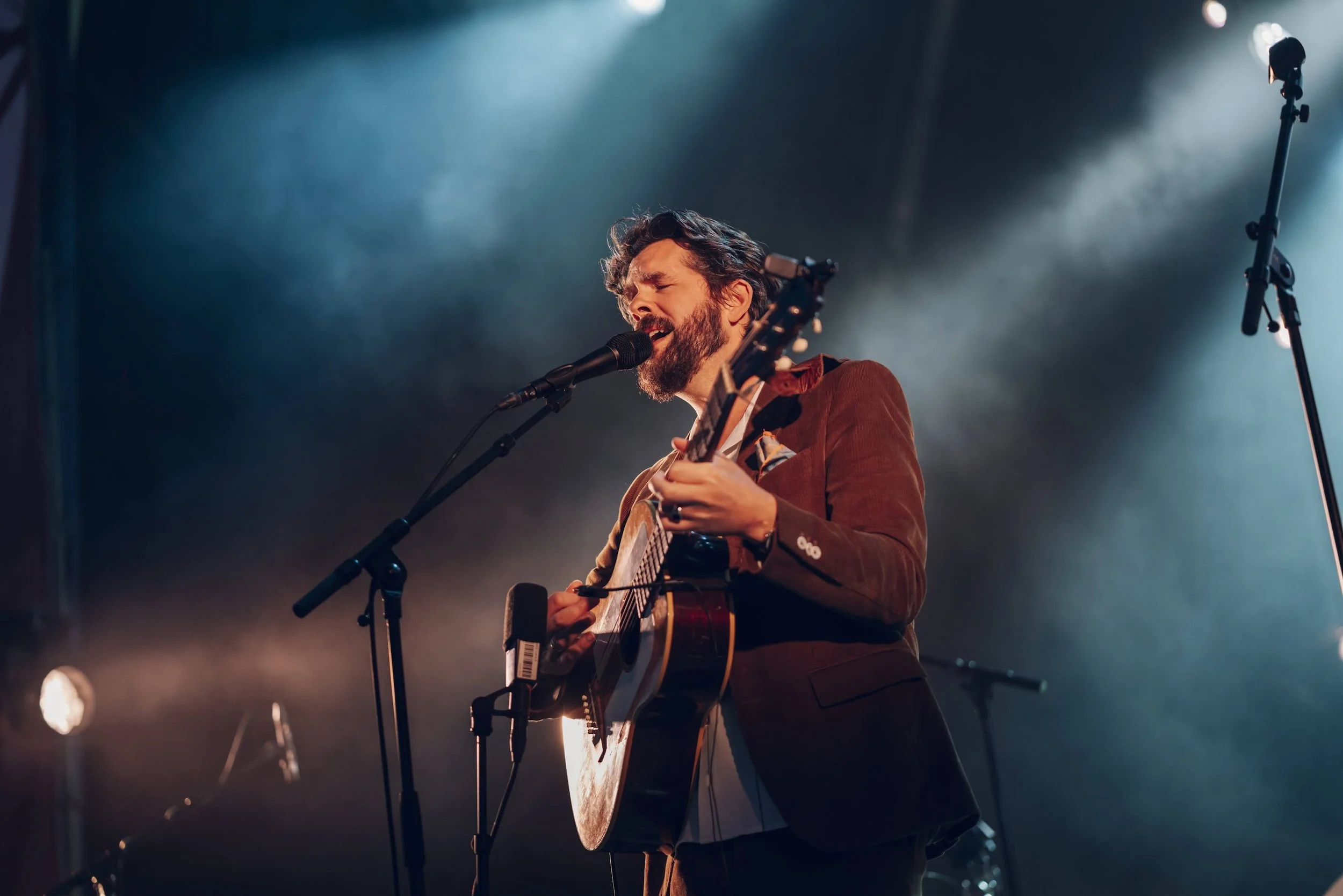 En mann som spiller akustisk gitar og synger inn i mikrofon på en scen under en konsert med blå og oransje lys.