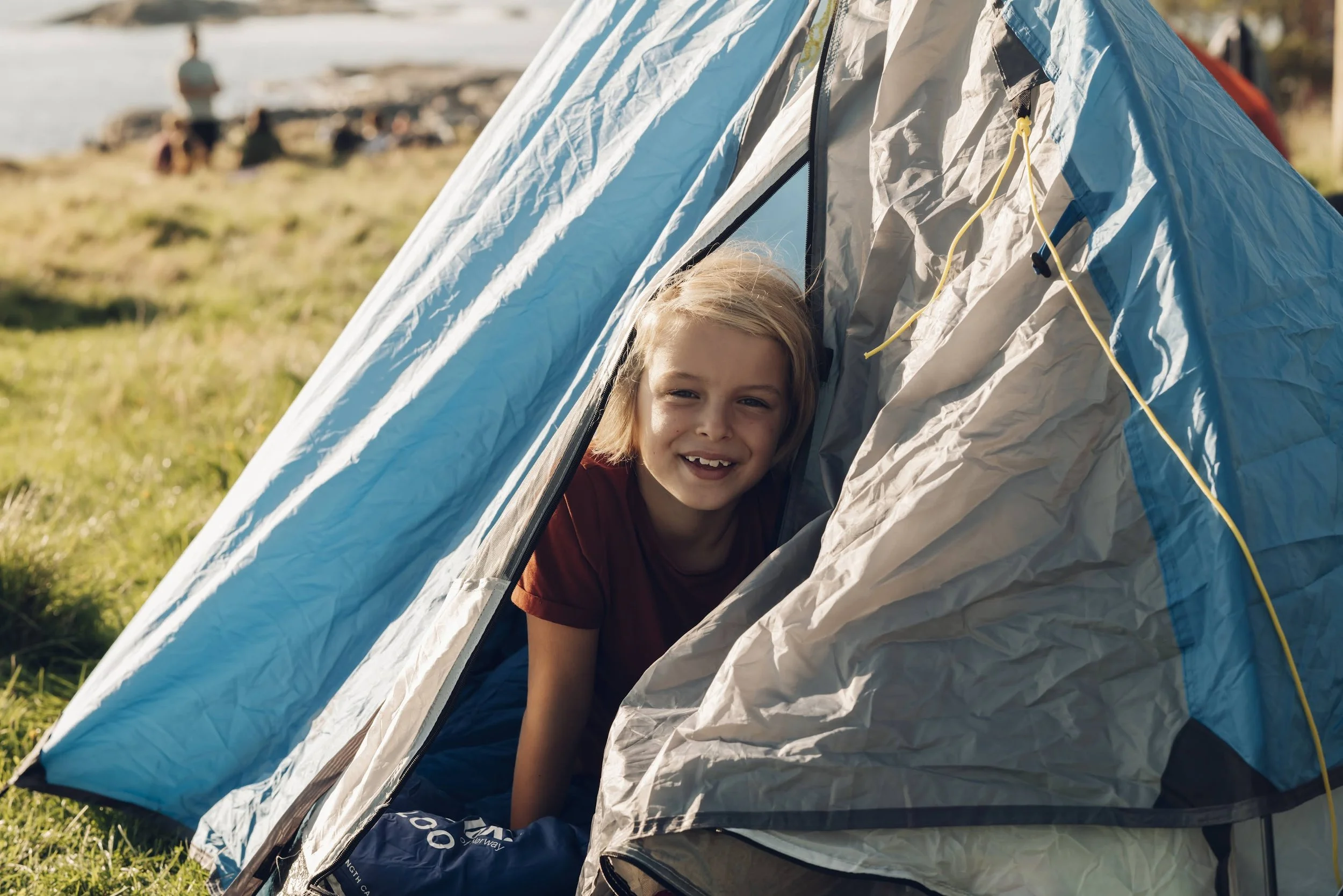 En ung jente kikker ut av en blå og grå telt på en campingplass ved vann. Hun smiler og har på seg en rød t-skjorte, med andre campere i bakgrunnen.