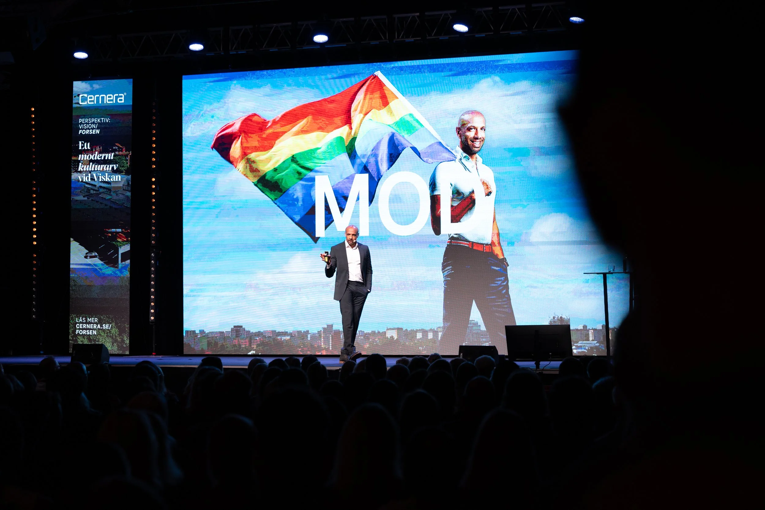Aaron Kroon står på en scen under en föreläsning, medett foto på en regnbågsflagga i bakgrunden och ordet ”mod” synligt bakom honom.