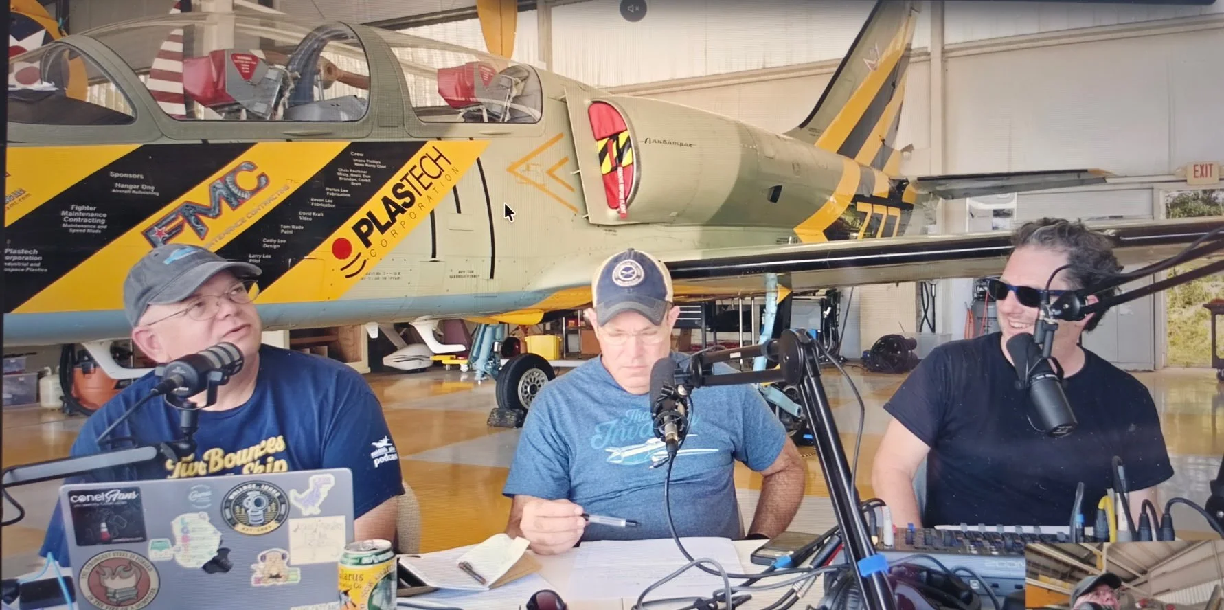 The three hosts on microphones at the live show, with an L-39 jet parked behind them