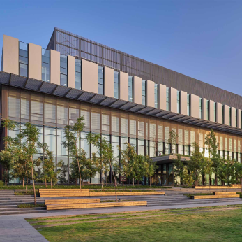 Modern multi-story building with large glass windows, surrounded by trees and benches on a sunny day.