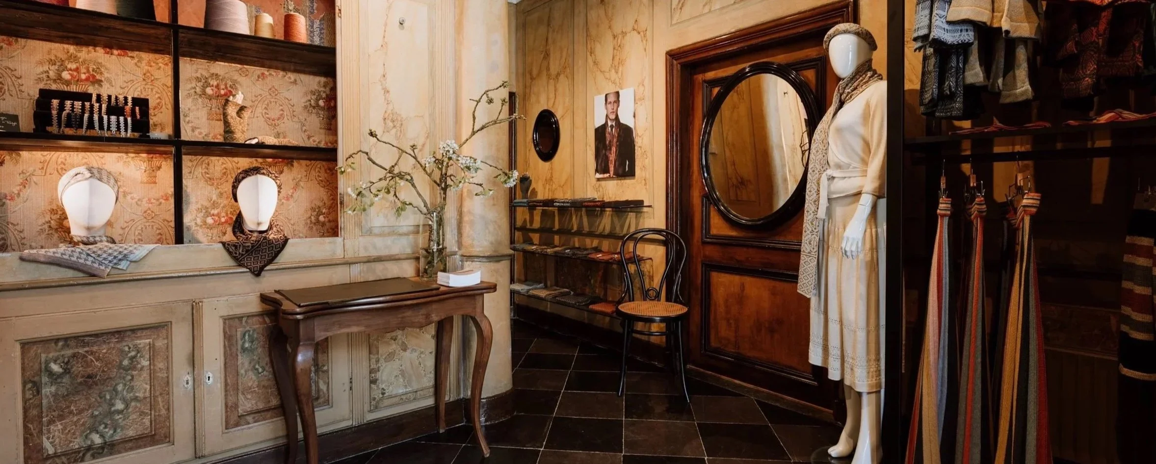 Interior of a boutique or clothing store with mannequins, display shelves with accessories, a small table, a chair, and a mannequin dressed in vintage-style clothing.