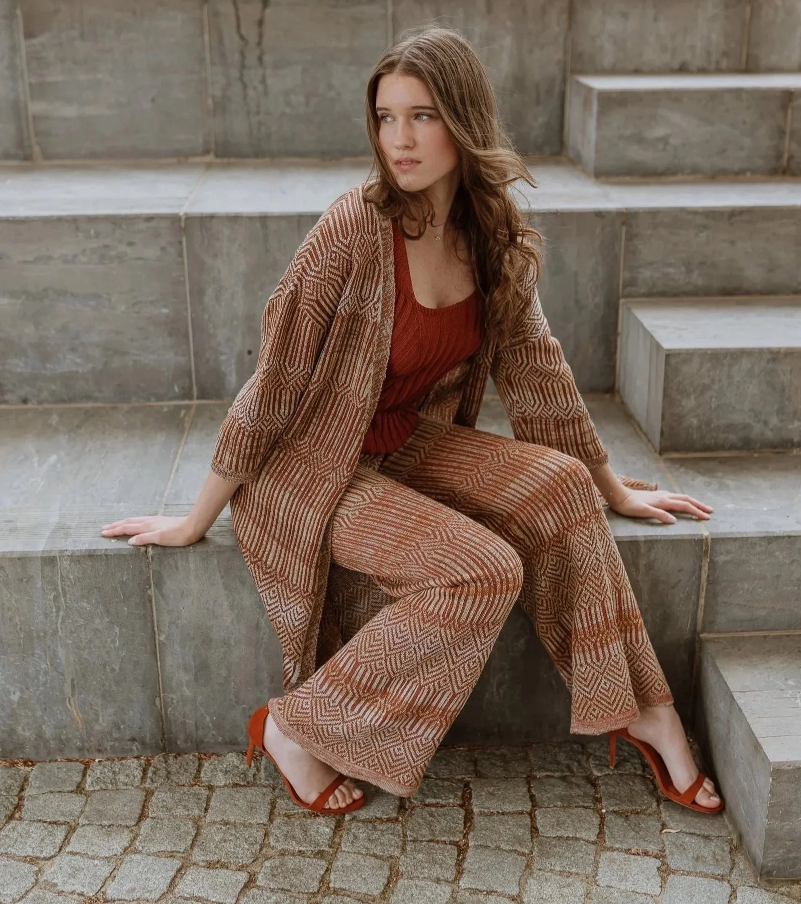 Model seated on stone architectural steps wearing a linen two-piece ensemble in warm earth tones, combining a patterned outer layer with a rust-coloured top.