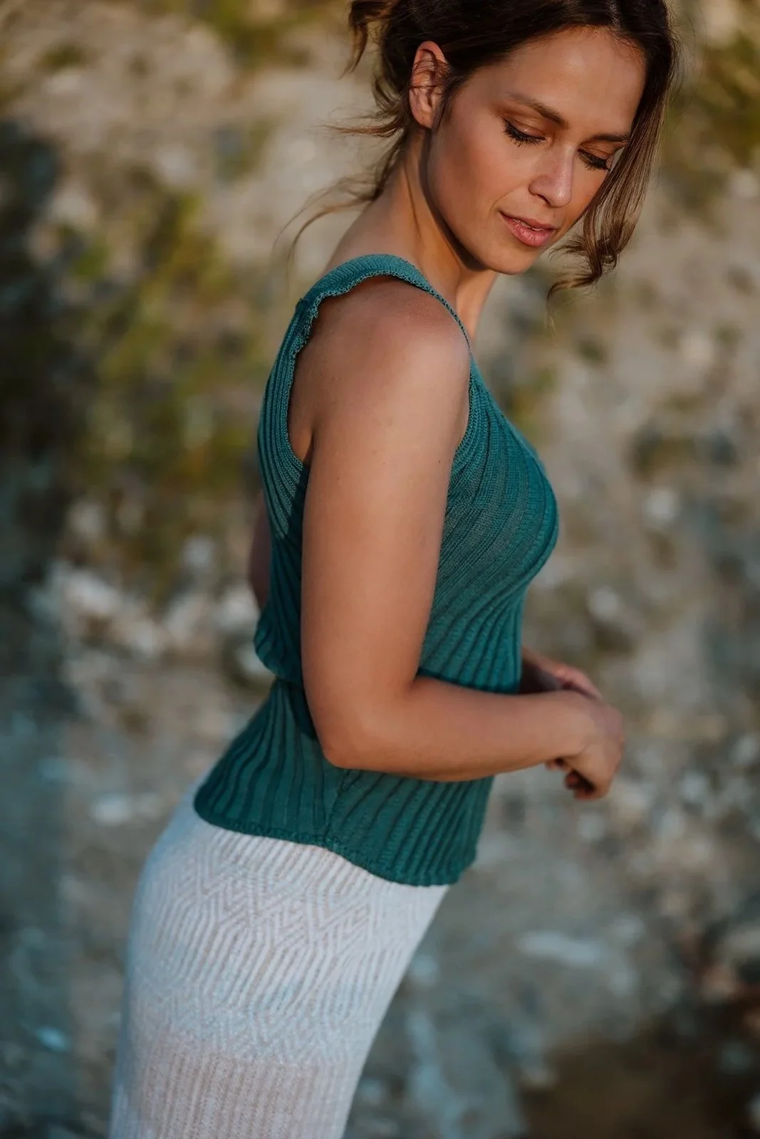 A woman in a teal sleeveless top and white patterned pants standing outdoors near water with her eyes closed and head tilted downward.