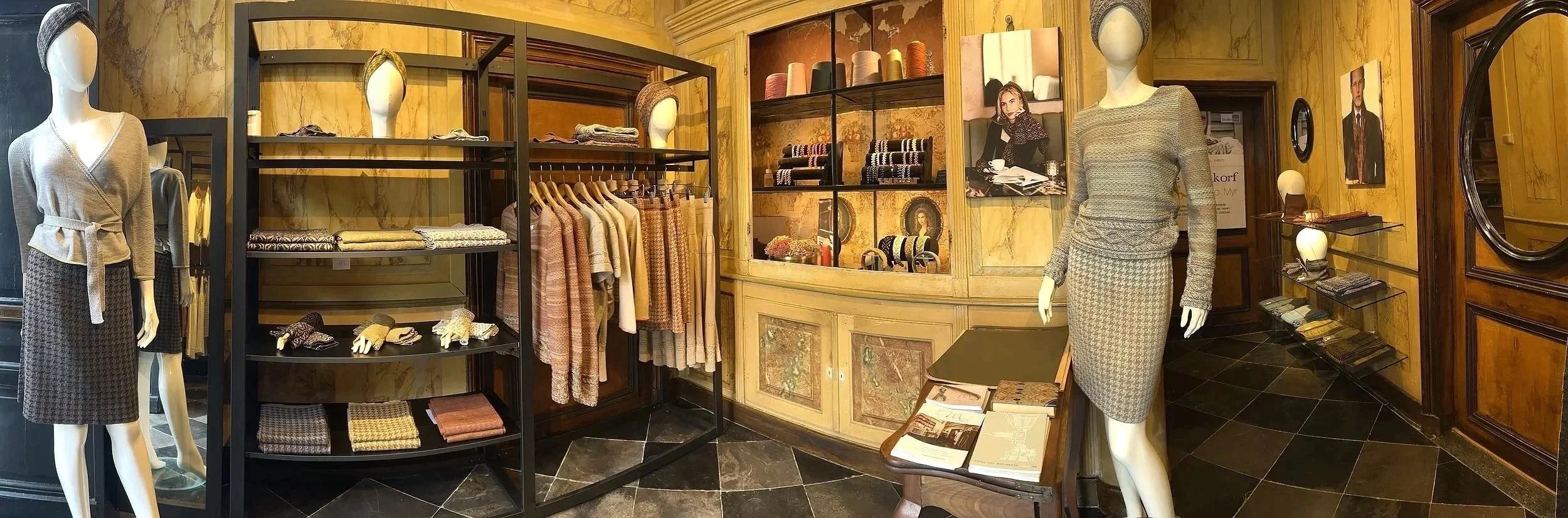 Mannequins dressed in beige and gray skirts and sweaters, surrounded by shelves and racks of folded clothes, socks, scarves, and accessories inside a boutique with yellow walls and black tiled floor.
