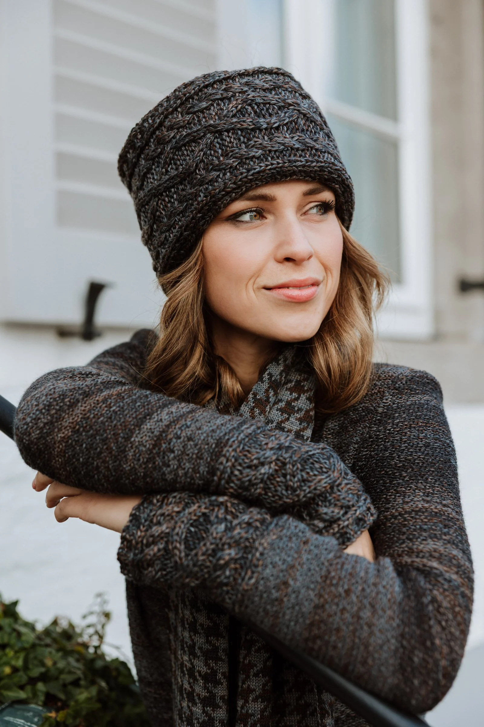 A woman with brown hair and blue eyes, wearing a dark knit hat and a cozy, multicolored sweater, is sitting outdoors near a white house with shutters, looking into the distance.