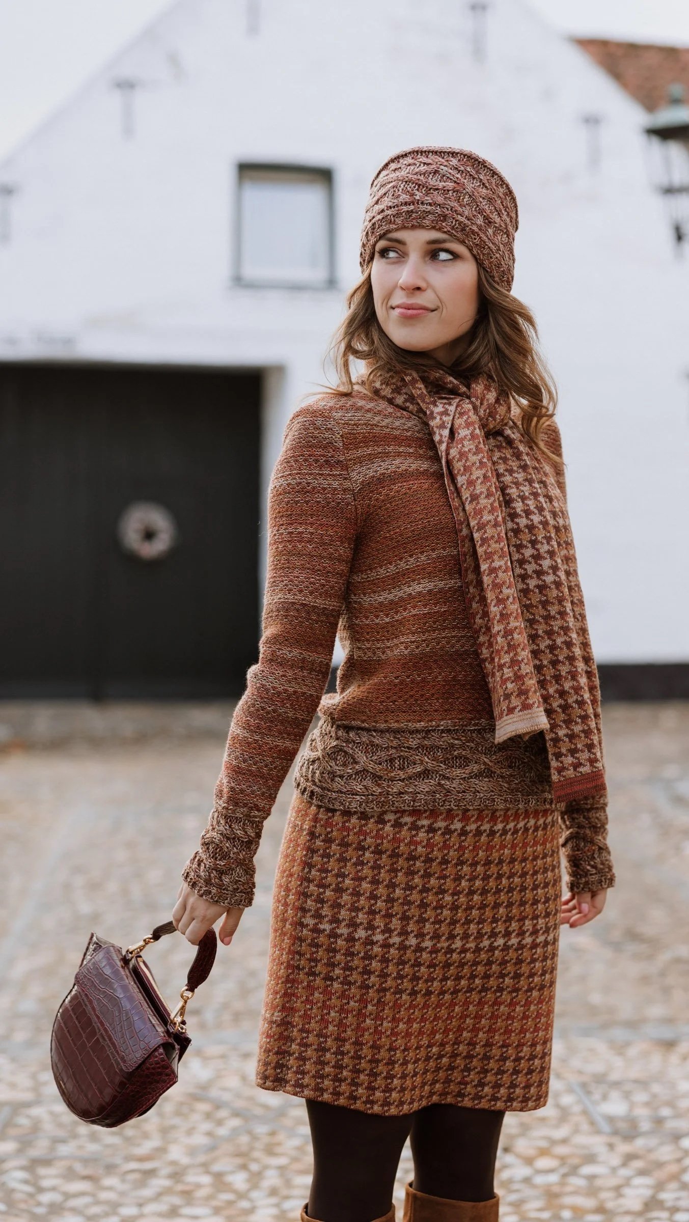 A woman dressed in warm autumn clothing including a knitted hat, scarf, and sweater standing outdoors near a white building with black doors.
