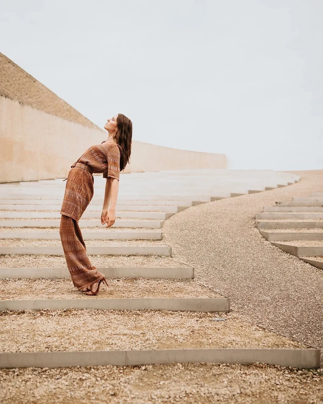Model wearing a linen jacquard two-piece set in warm earth tones, standing on architectural stone steps in an open landscape.
