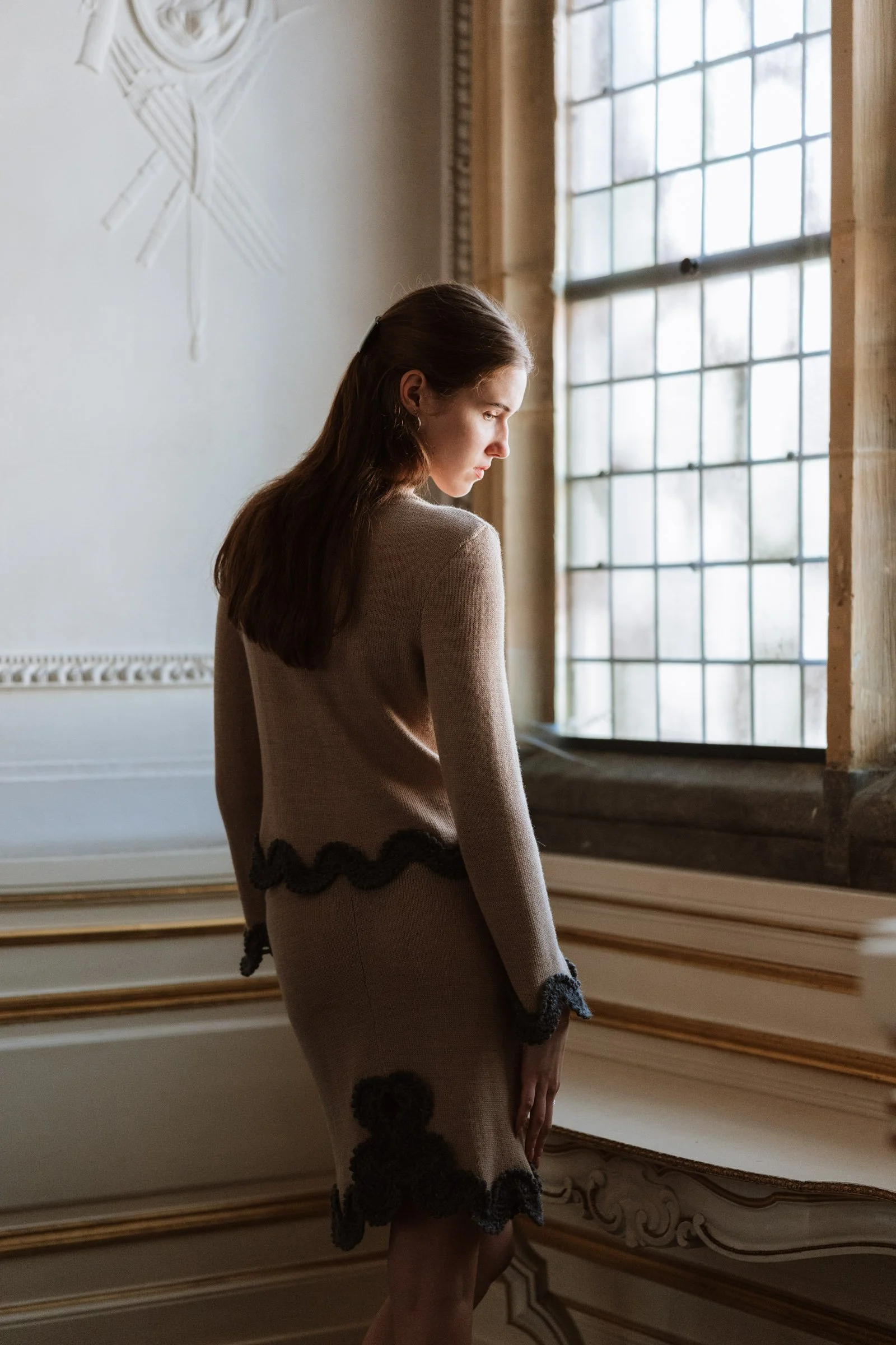 Studio Myr BAROQUE – Back view of the Gabriella jumper in beige Merino wool with grey crochet lace edging, worn by a window