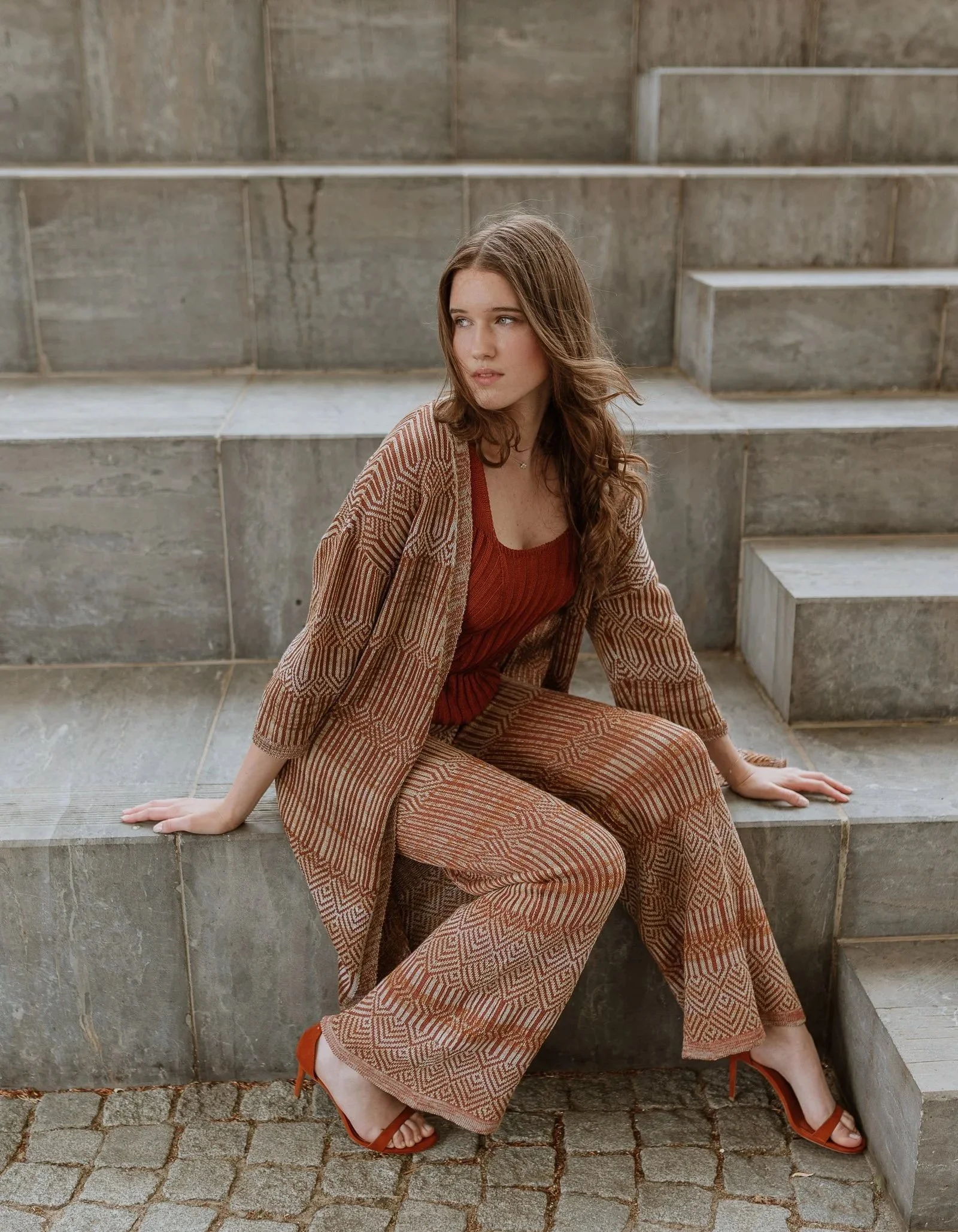 Woman wearing a refined jacquard knit summer ensemble from the Studio Myr African Nomads collection, featuring warm earthy tones and geometric patterns, photographed on architectural stone steps in soft natural light.