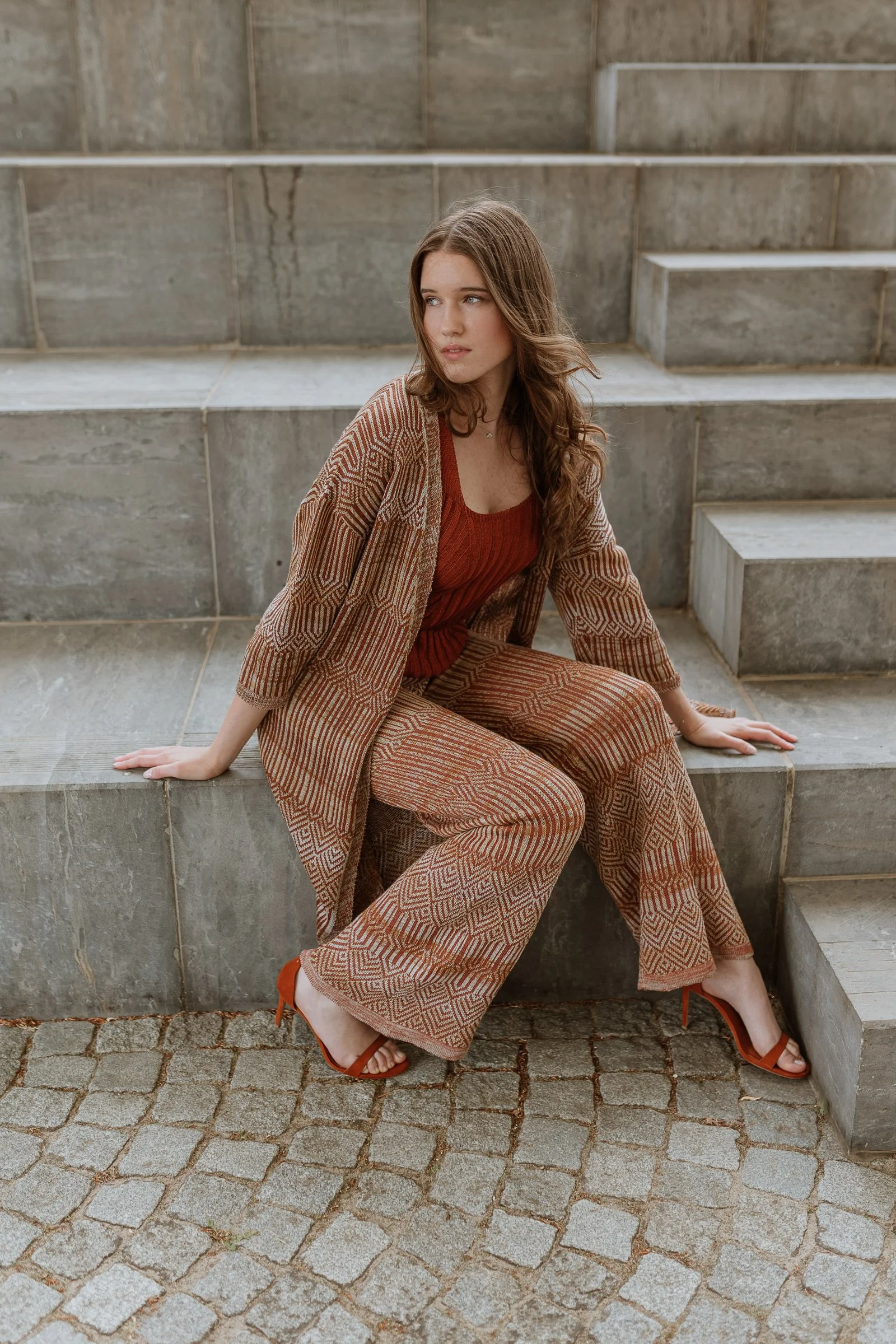 A young woman with long brown hair sitting on stone steps outdoors wearing a rust-colored top, patterned wide-leg pants, a matching patterned blazer, and rust-colored high-heeled sandals.