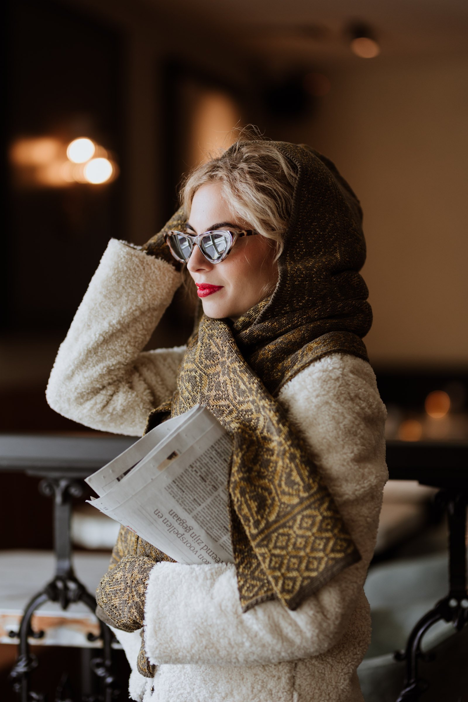 A woman with curly blonde hair wearing sunglasses, a patterned headscarf, a warm coat, and patterned gloves, holding a newspaper in a cozy indoor setting.