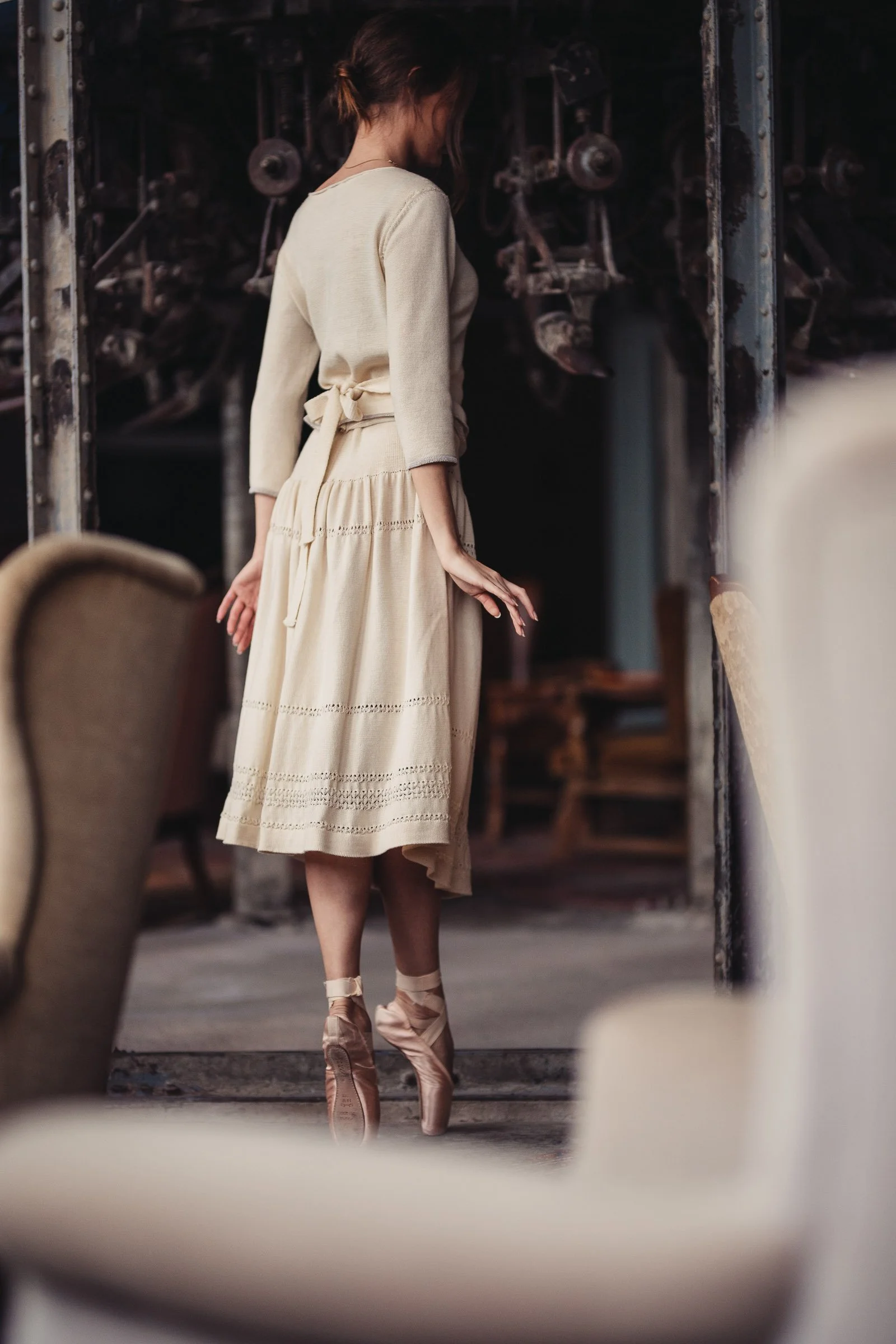 A woman dressed in a beige cardigan and skirt standing on pointe ballet shoes in an industrial setting.