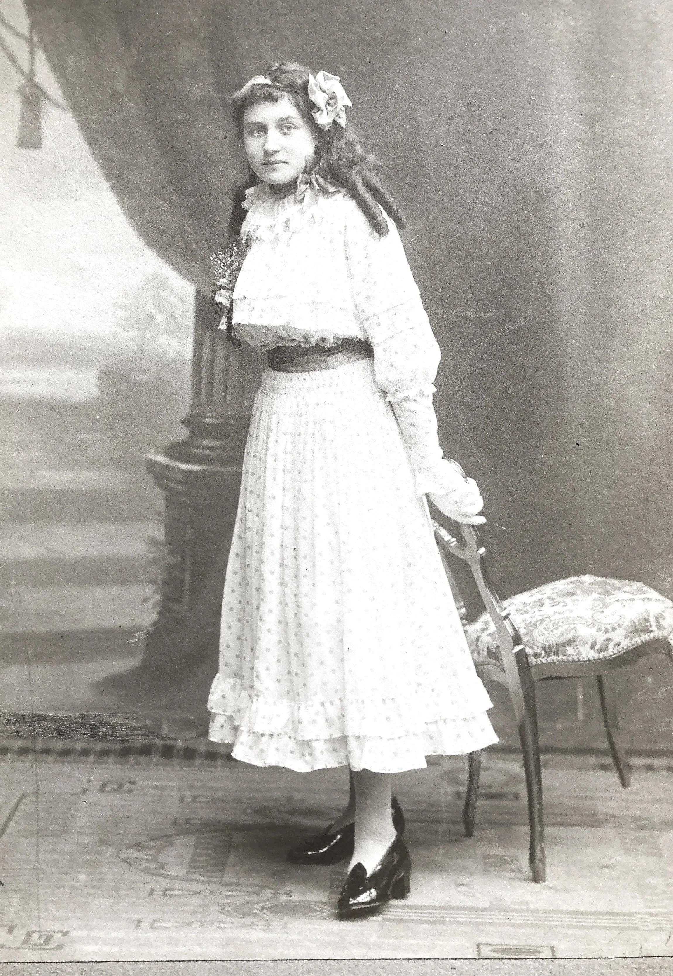 Archival black-and-white portrait of the founder’s grandmother as a young girl, circa 1915, wearing a light dress and standing in a studio setting.