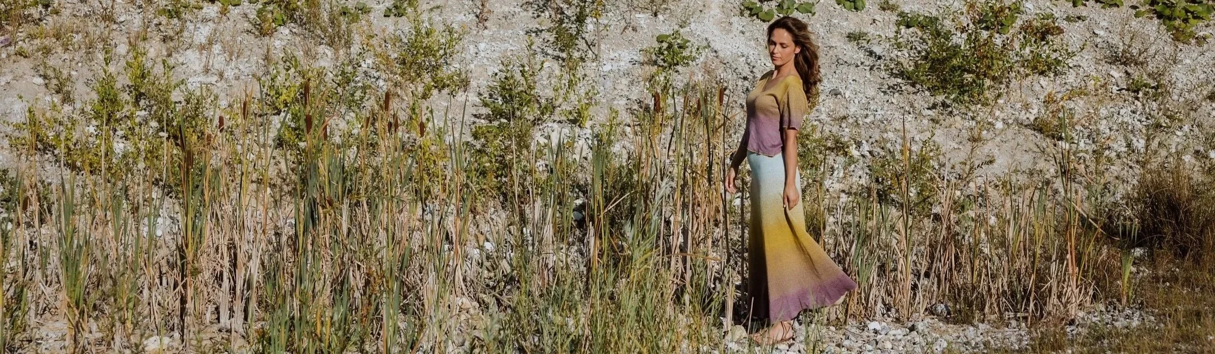 A woman standing among tall grass and bushes in a rocky outdoor setting, wearing a multicolored dress with shades of yellow, brown, purple, and blue.