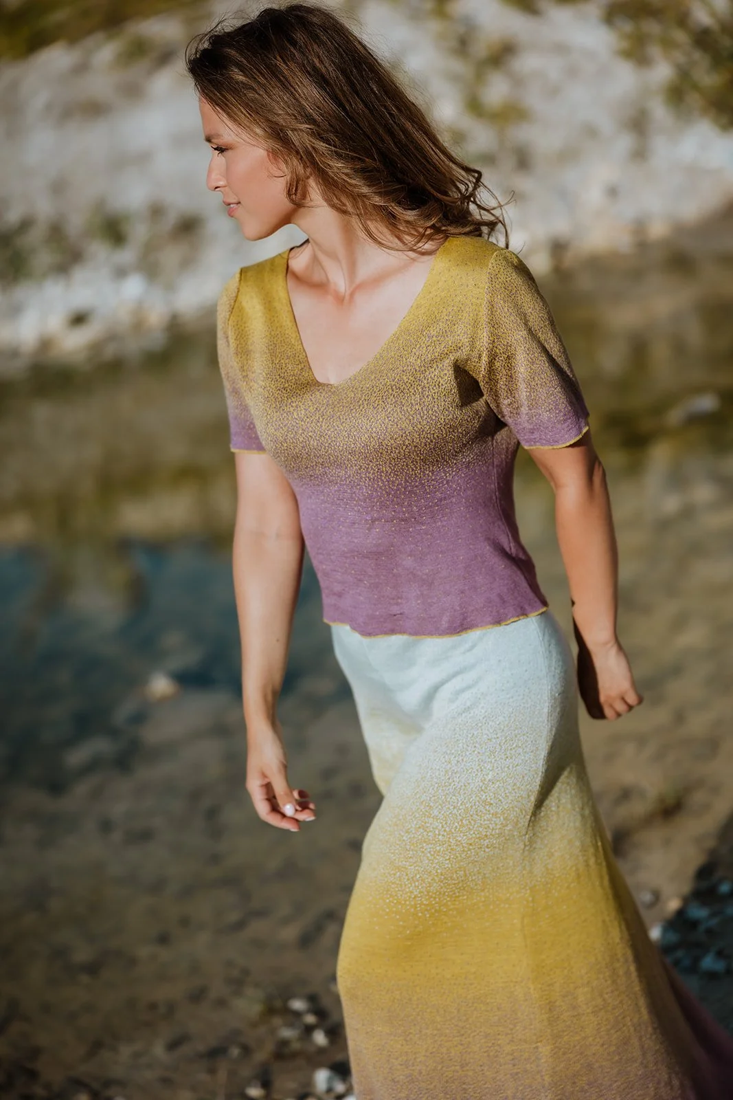A woman is walking along a rocky shoreline, wearing a gradient-colored dress that transitions from purple to yellow, with a V-neck and short sleeves, her long hair flowing behind her.