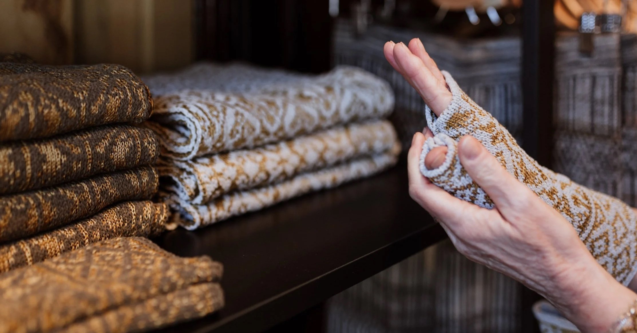 Close-up of a person's hand wearing a fingerless glove, folding a patterned textile on a dark shelf, with neatly folded similar textiles in the background.
