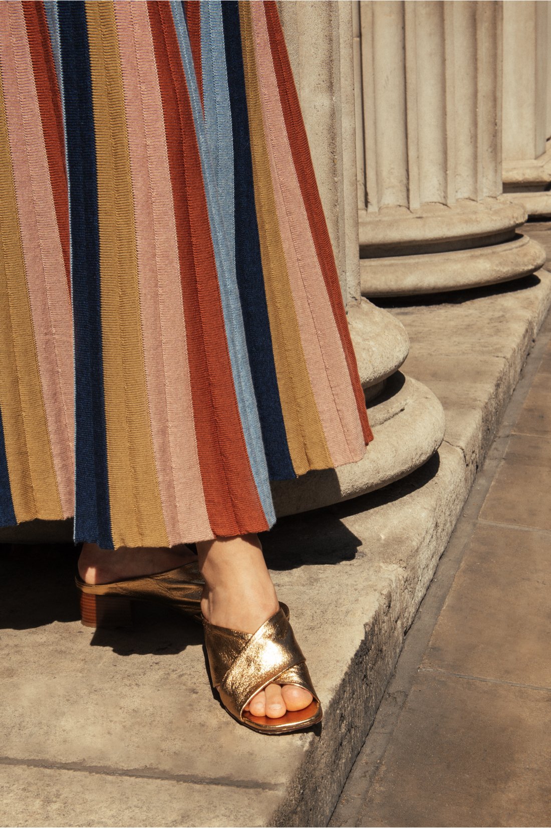 Close-up of Studio Myr knitted skirt with multicolour stripe pattern, worn with elegant footwear against a classical stone backdrop.