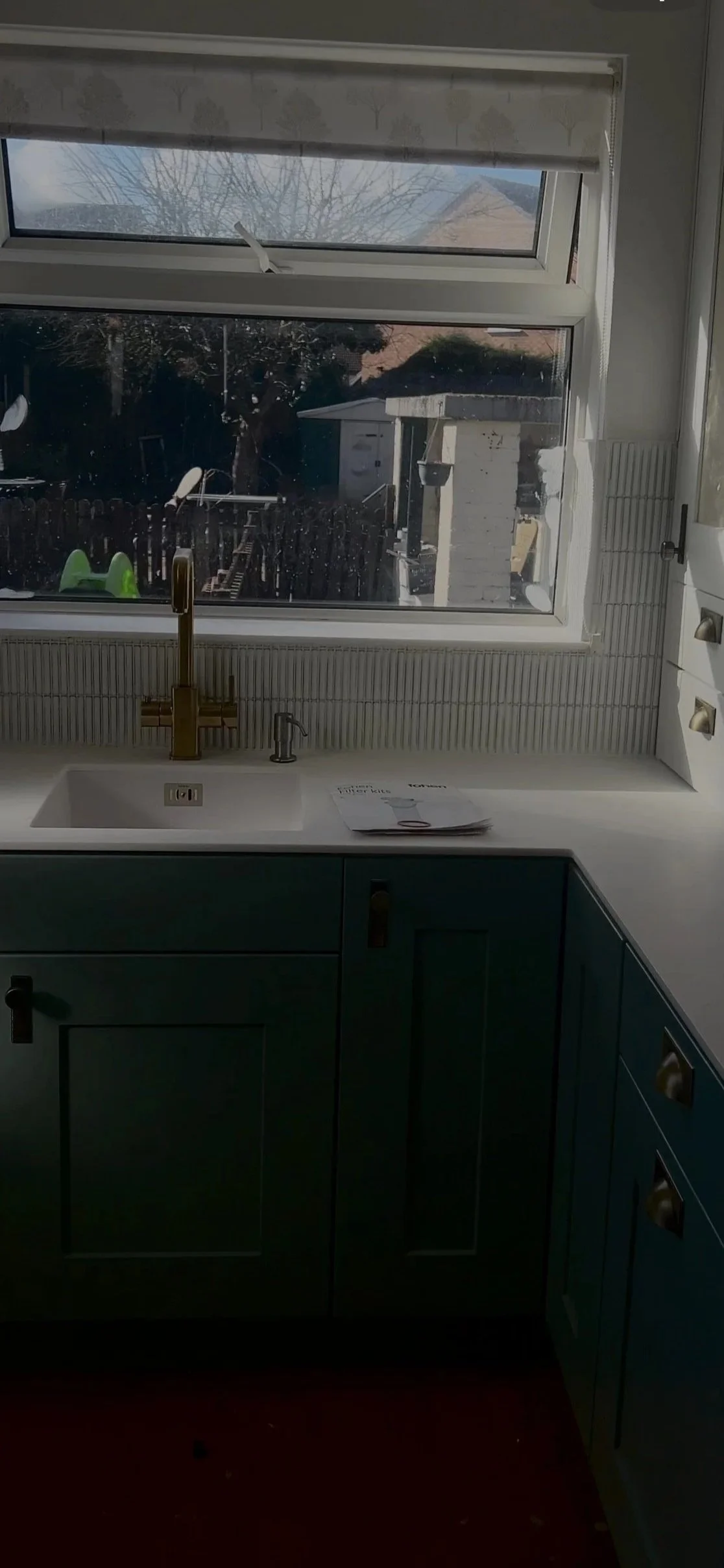 A kitchen with a window showing a backyard, a sink with a gold-tone faucet, and blue cabinetry beneath the countertop.