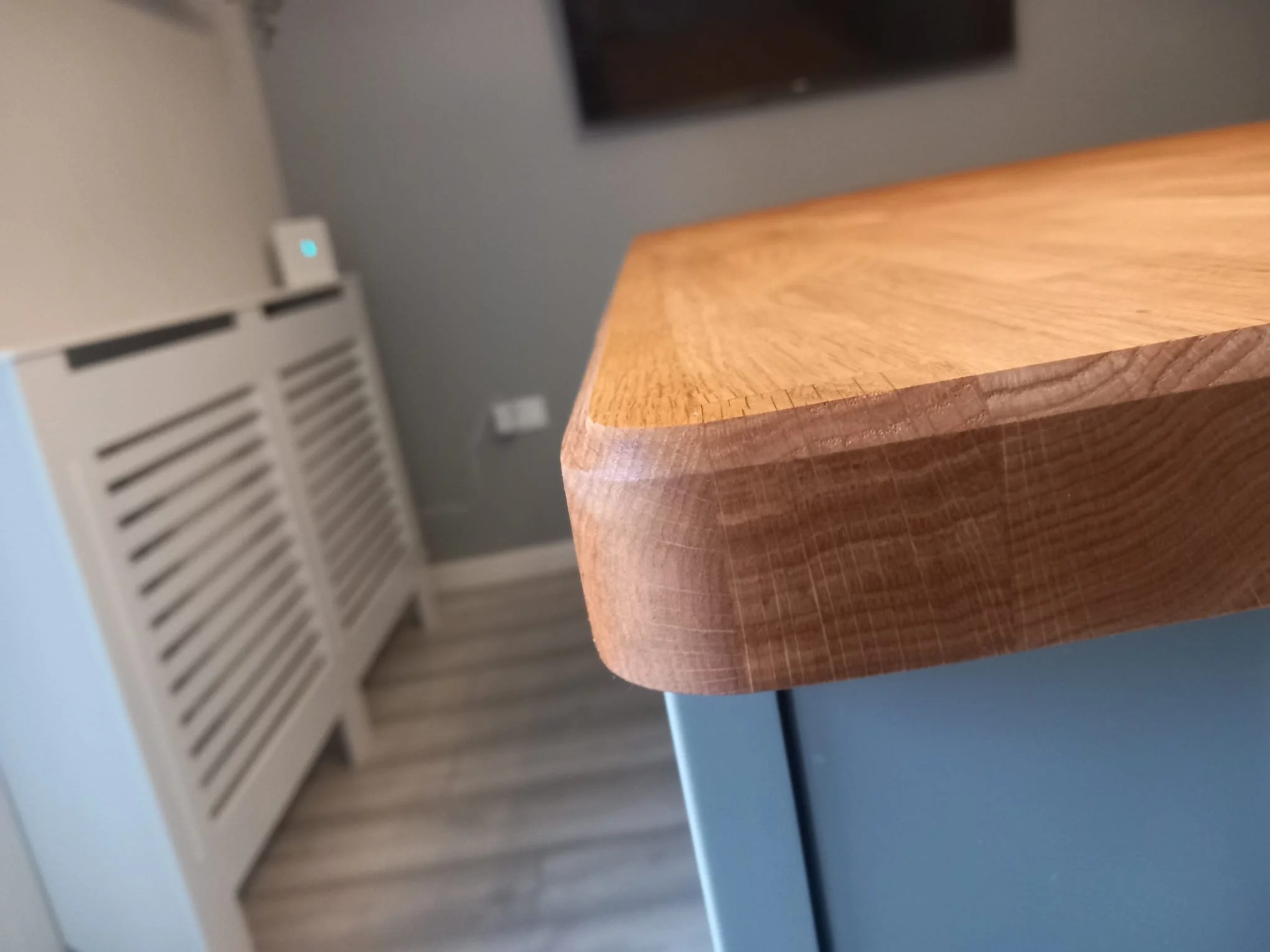 Close-up of a wooden table edge with a background of a wall, vent, and some objects in a room. Worktop fit, Kitchen fit, Vertex solid surface 