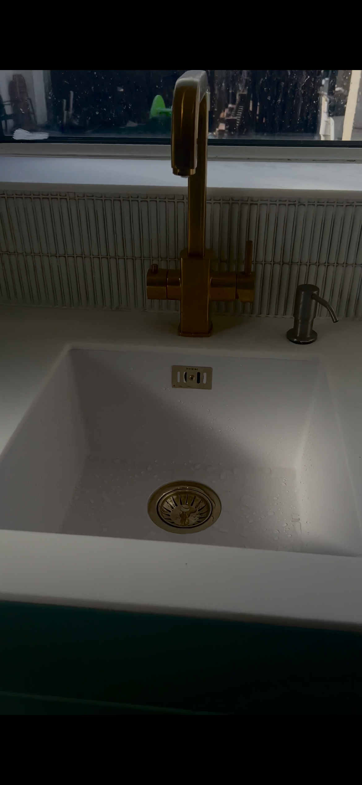 White kitchen sink with a gold faucet, a small gray soap dispenser, and a window above showing a cityscape outside.