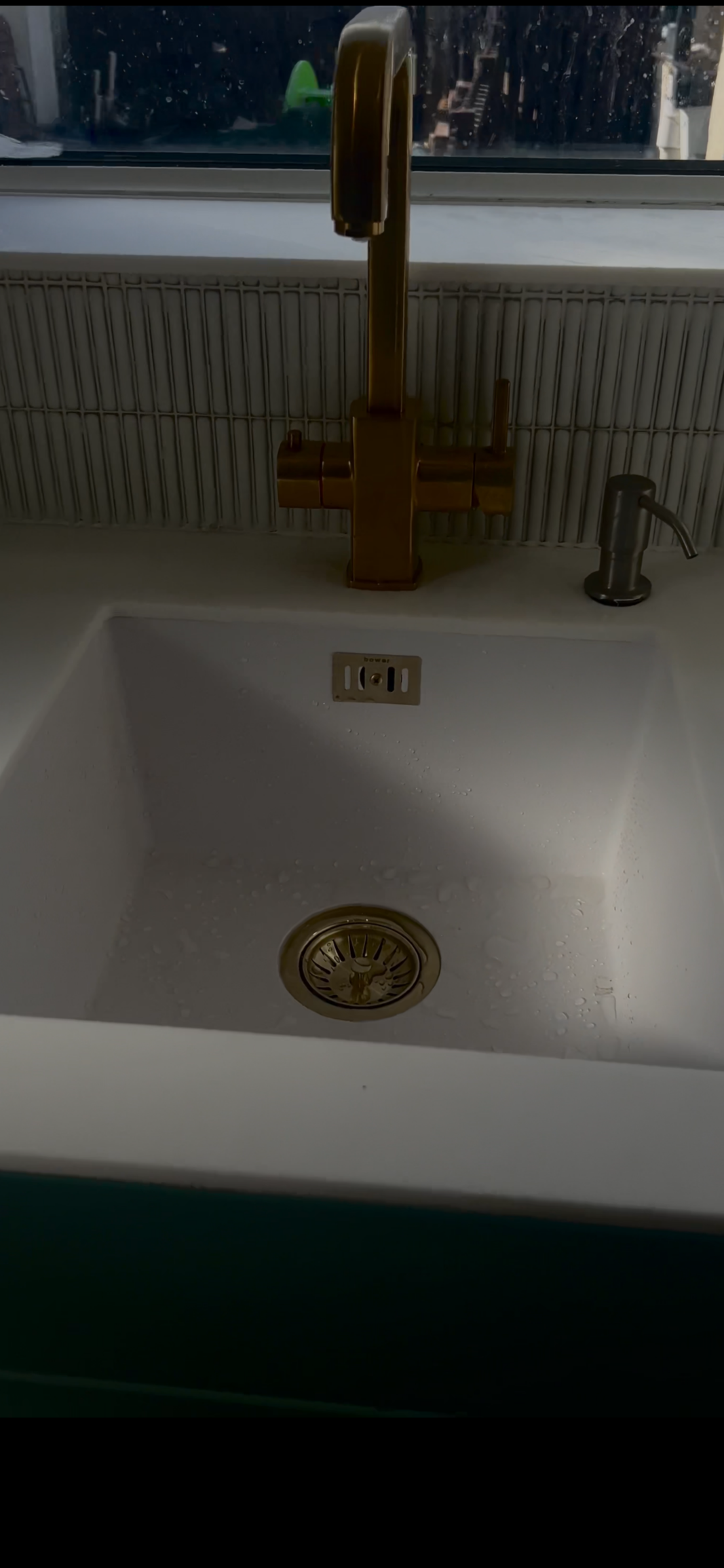 Kitchen sink with a gold-colored faucet, a small silver soap dispenser on the right, and a window above showing rain outside.