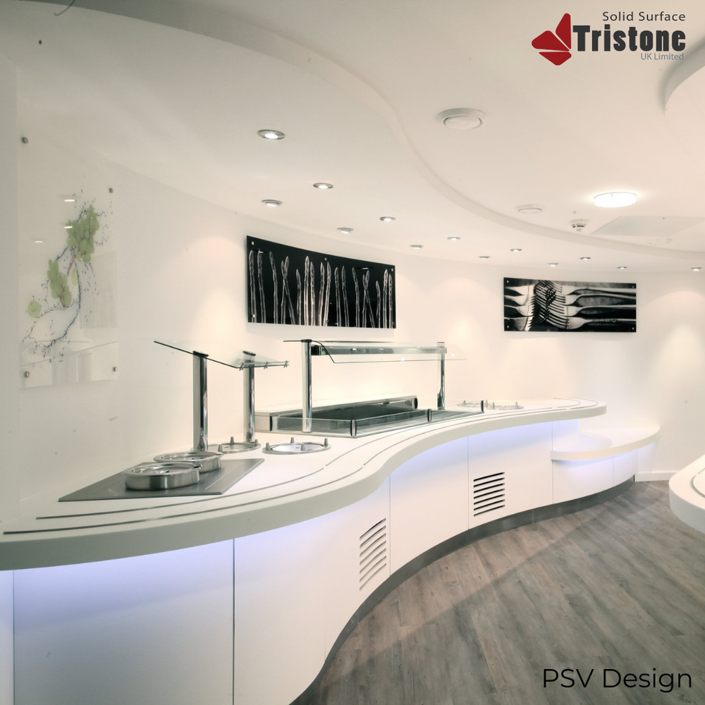 Modern buffet area with white curved counter, abstract artwork on white walls, and metal food covers, in a well-lit setting.