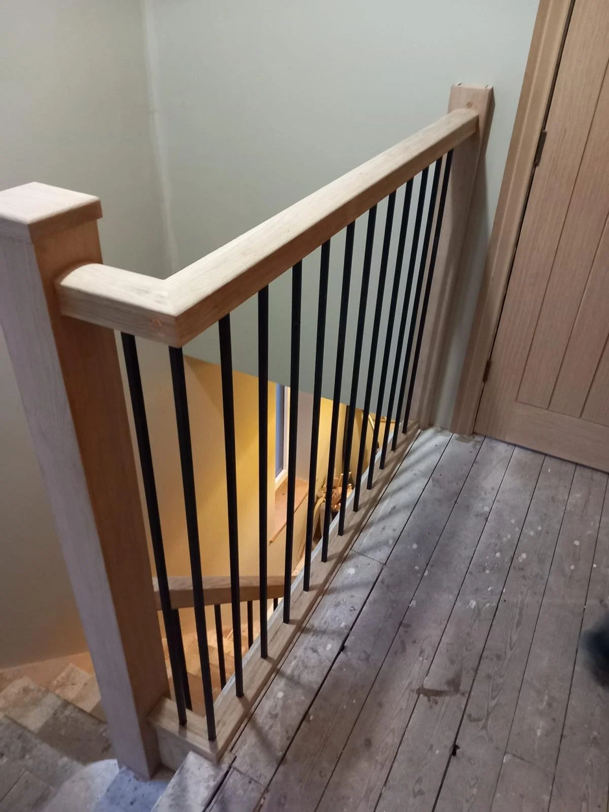 Indoor wooden staircase with a wooden handrail and black metal balusters, next to a wooden door and wooden flooring.
