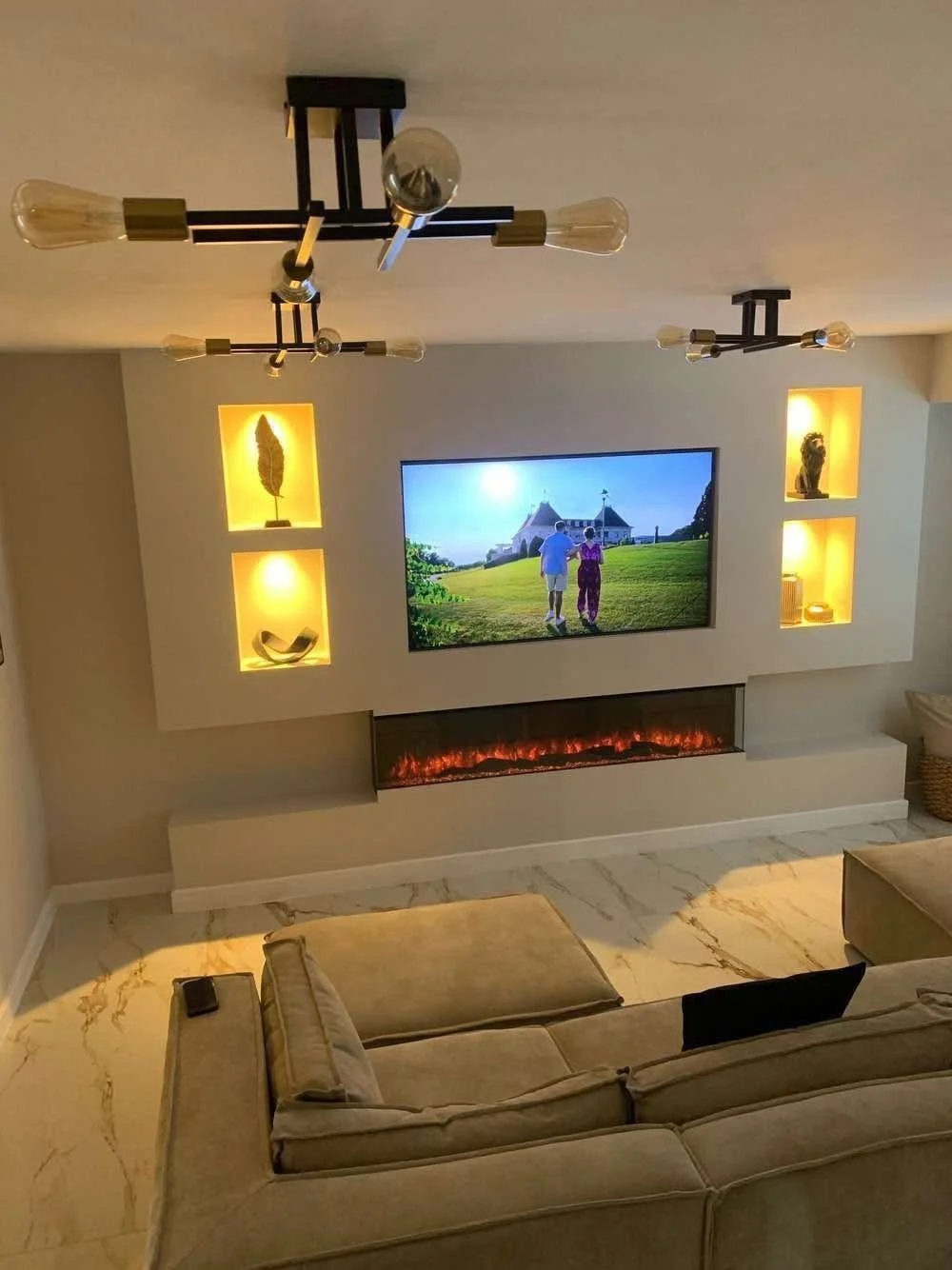 Living room with beige couch, wall-mounted TV showing people walking outside, electric fireplace beneath TV, and decorative shelves with sculptures and books. Modern ceiling light fixtures.