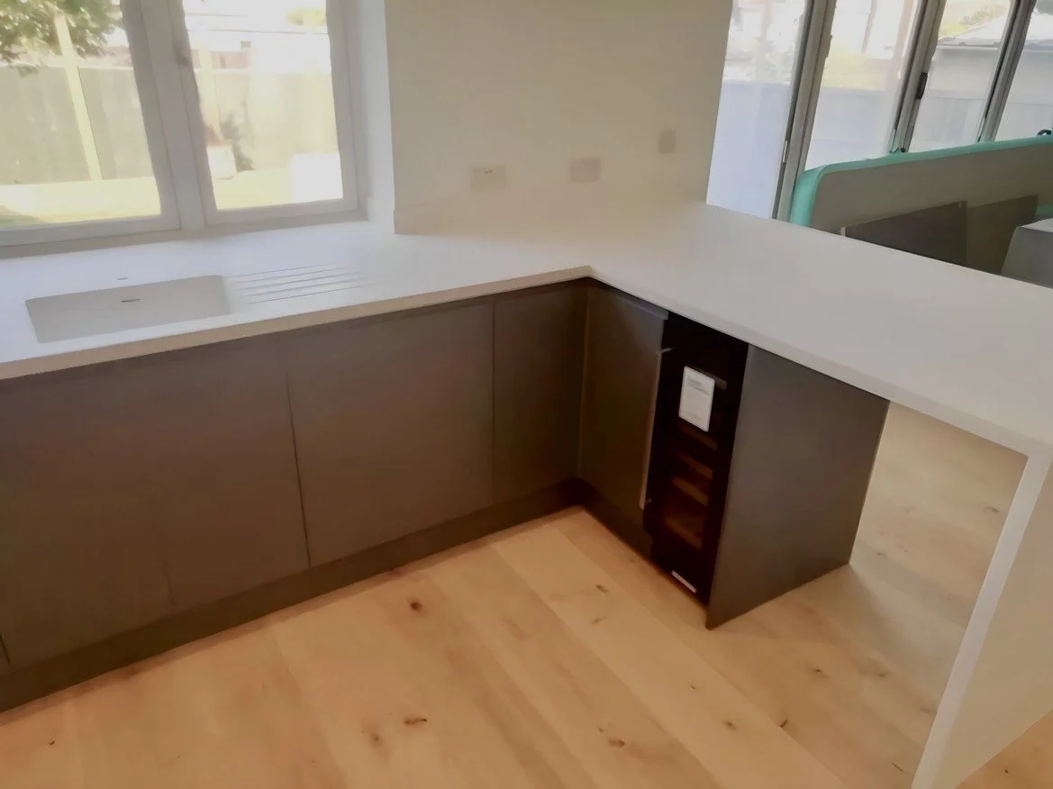 Empty kitchen corner with brown cabinets, white countertop, a window with a view of a yard, and a wine cooler under the counter.