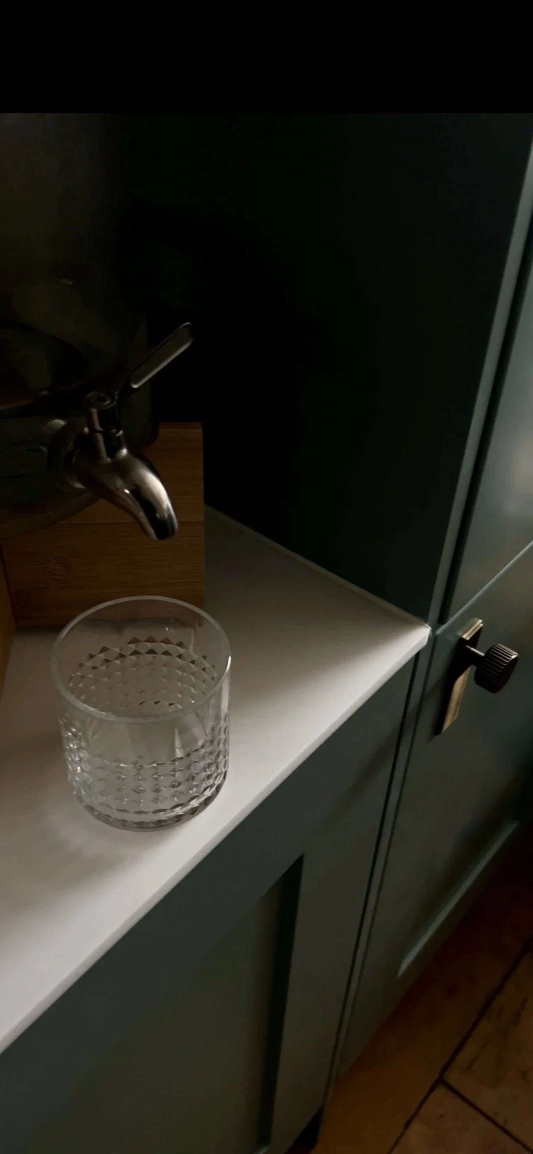 A glass of water on a white countertop near a kitchen sink with a silver faucet, adjacent to a cabinet with a dark gray door.