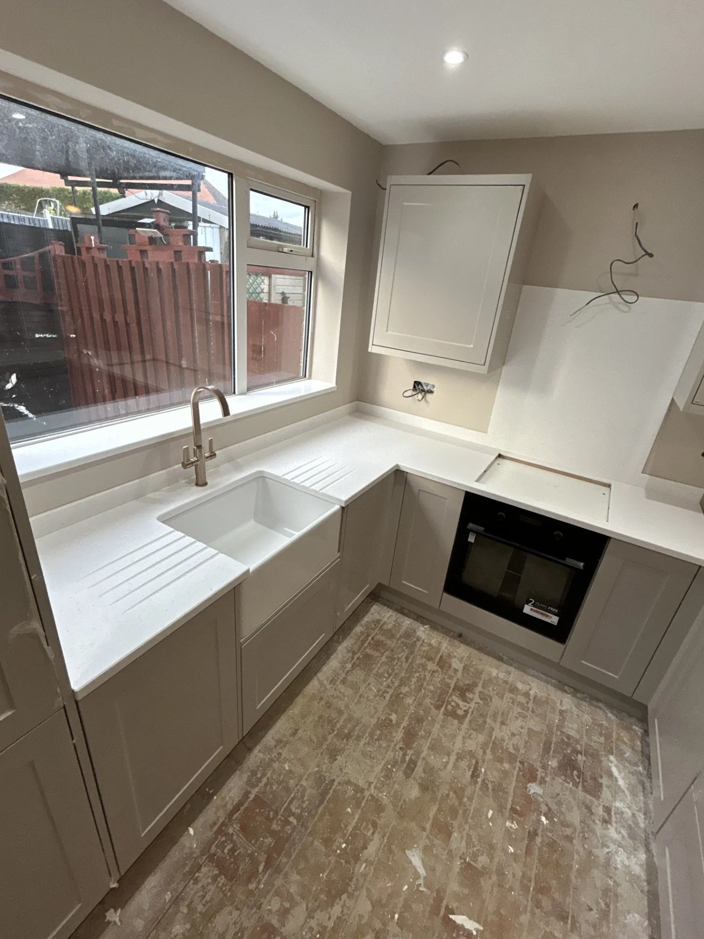 A kitchen under construction with white cabinetry, a built-in oven, a sink, and a large window overlooking a backyard with a red fence. Exposed wiring and unfinished surfaces are present.