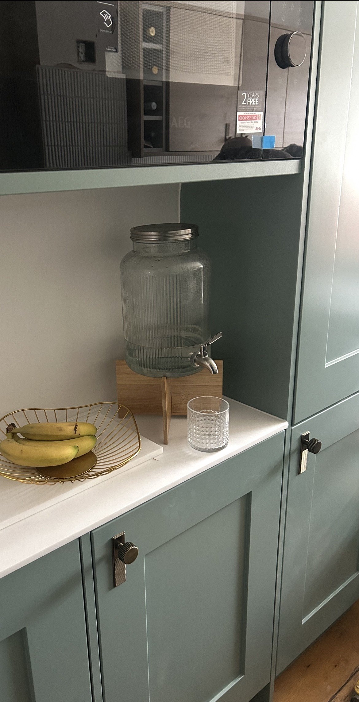 Glass water dispenser with a metal spigot on a wooden stand, a glass of water, a bunch o f bananas on a decorative wire tray, against a light-colored cabinet with black knobs, in a kitchen setting. joiner