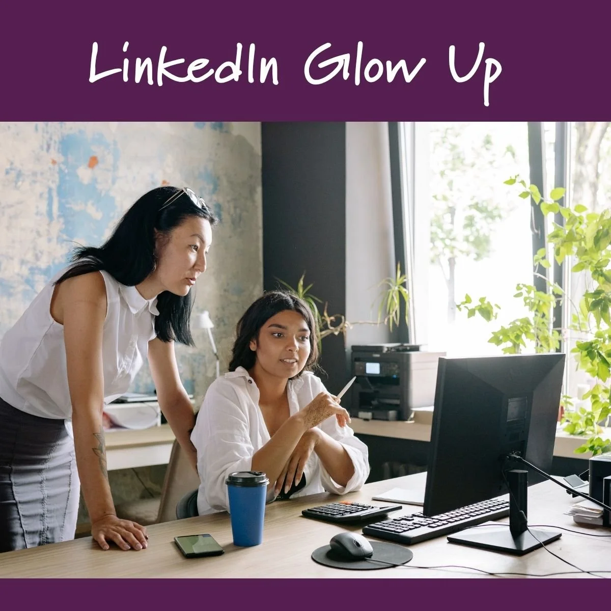 Two women working together in an office, one sitting at a desk and the other leaning over to look at the computer screen, with a coffee mug, phone, and keyboard on the desk.