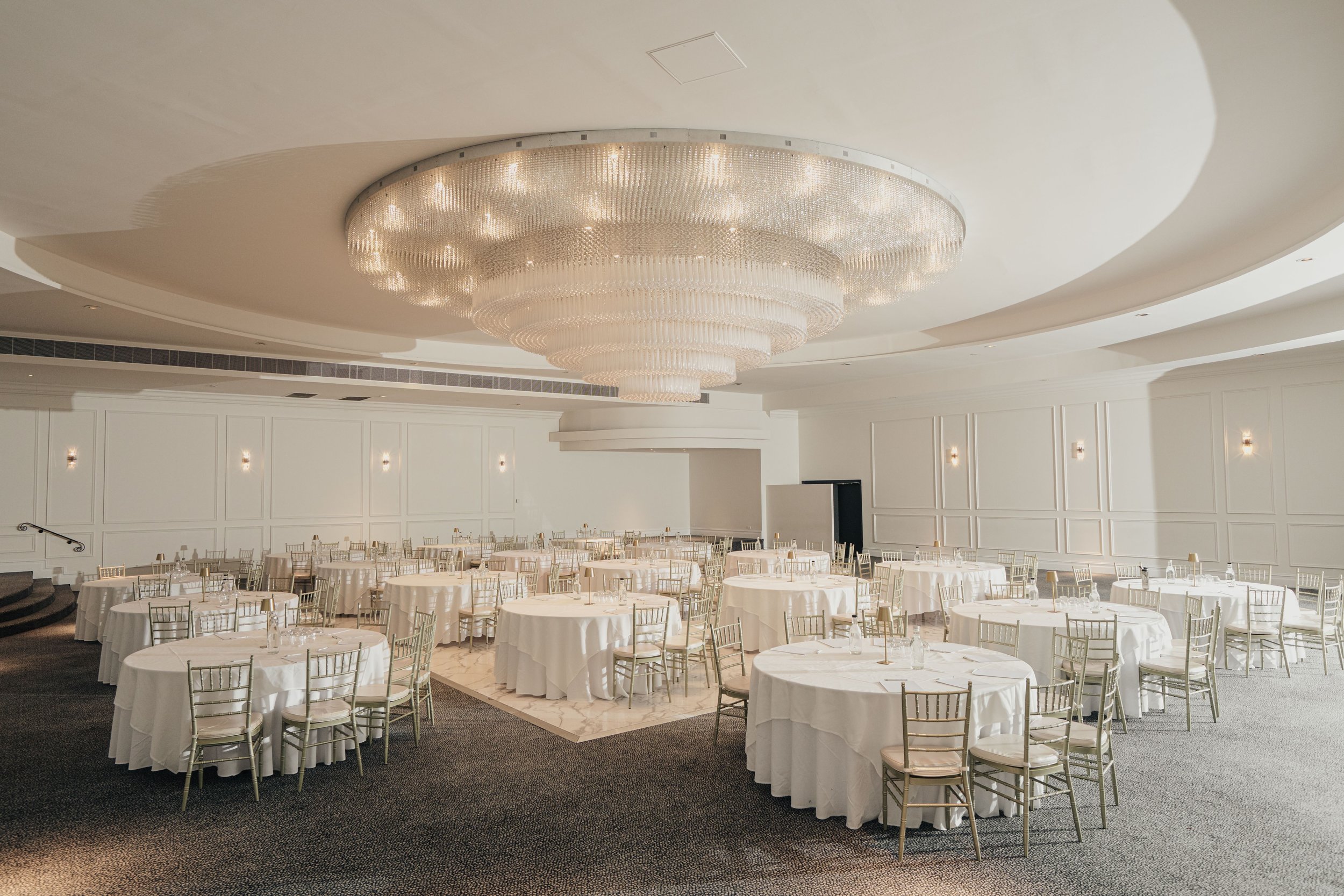 A bride and groom dancing in a large, elegant event hall with a grand chandelier and decorated tables.