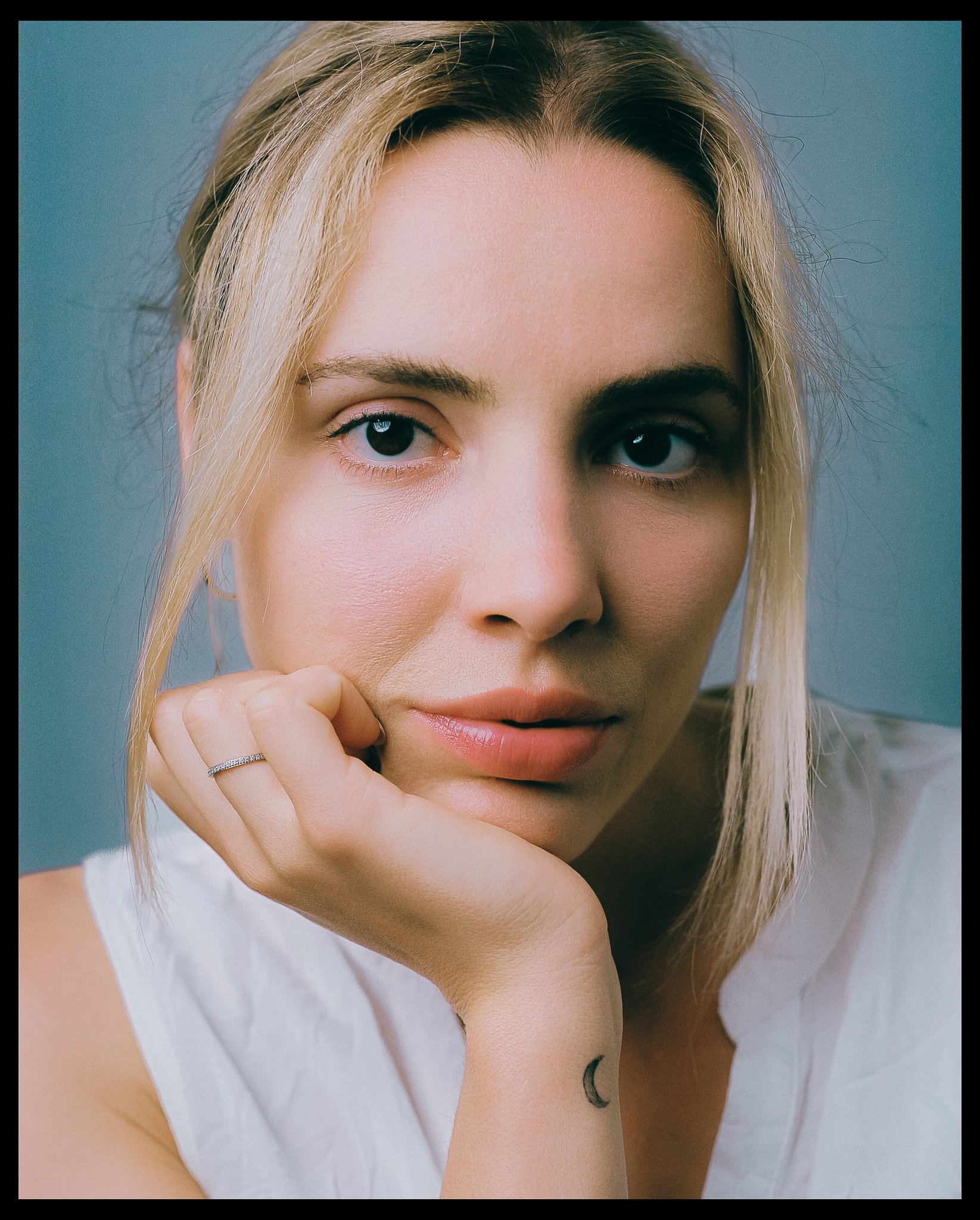 Mulher de cabelo loiro, expressão contemplativa, apoiando o rosto na mão, com fundo azul