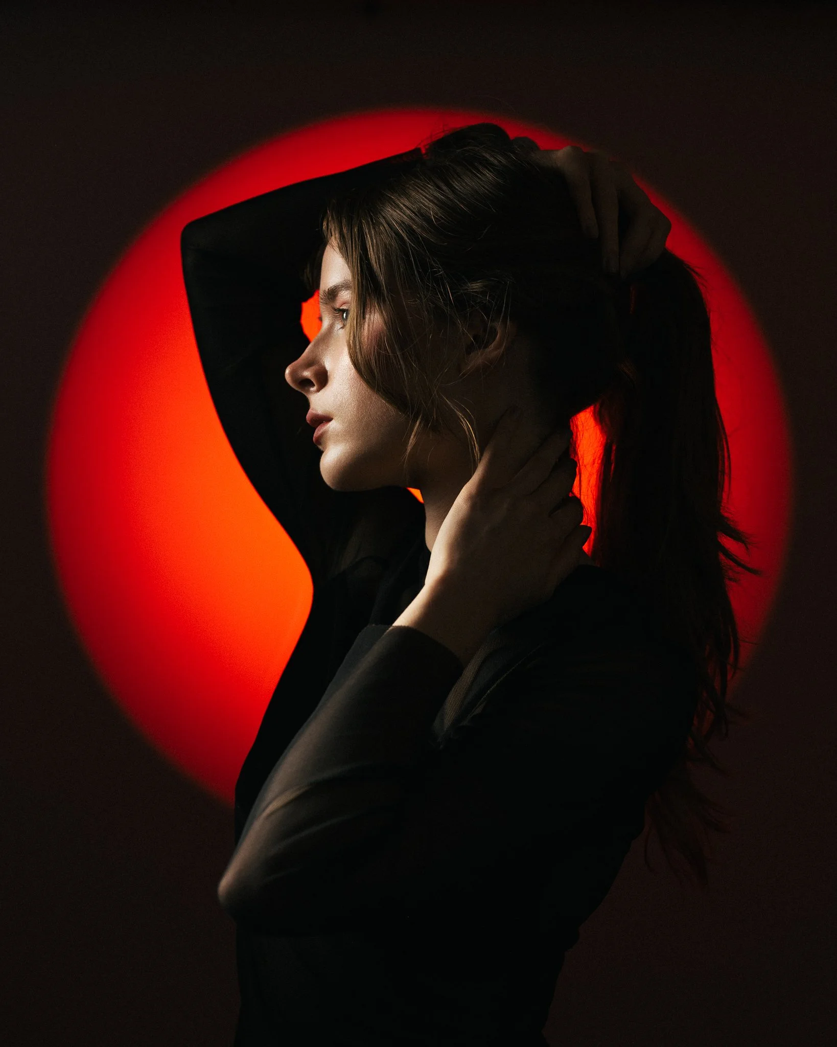 Retrato de uma mulher com cabelo castanho escuro, usando uma blusa preta, segurando a cabeça com uma mão, com fundo preto e uma luz vermelha circular atrás dela, criando um efeito dramático.