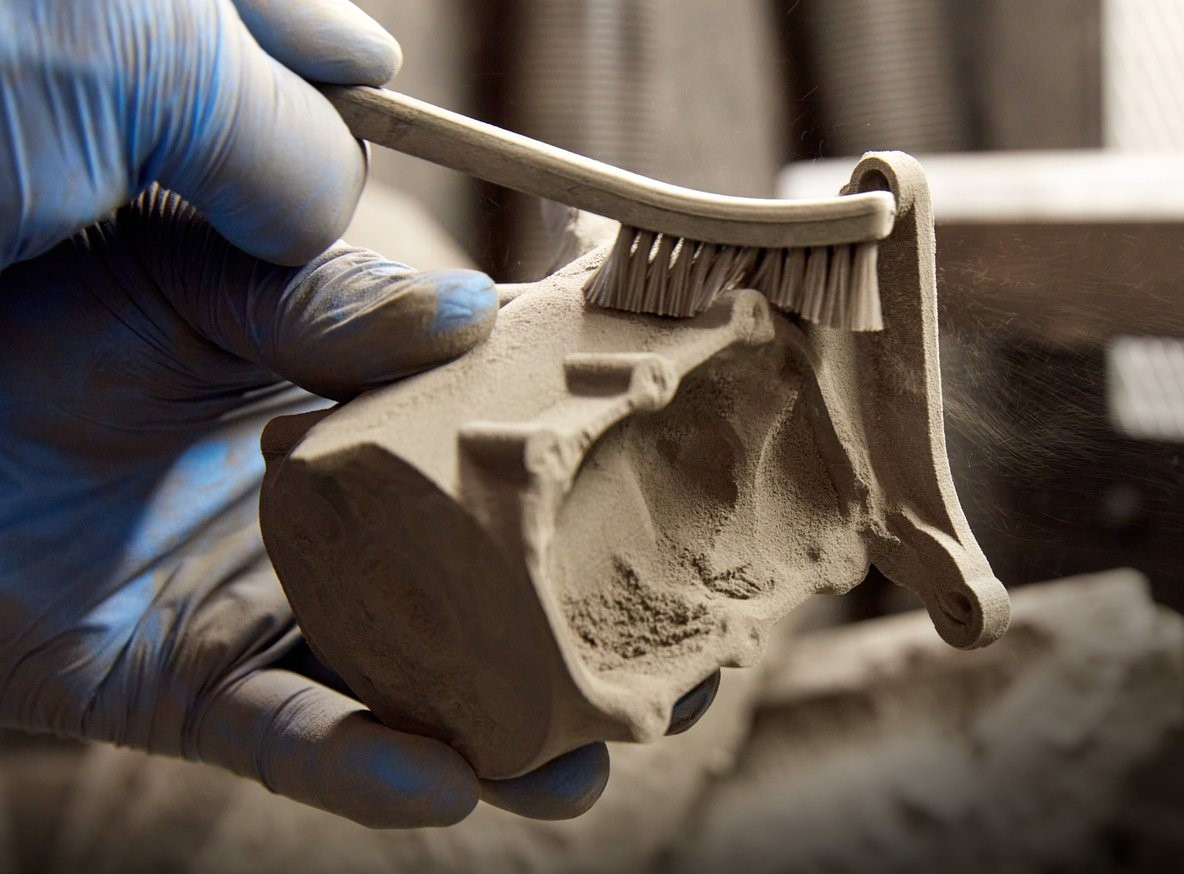 Handskbeklädd hand som borstar bort överflödigt pulver från en 3D-printad del under efterbearbetning för att illustrera pulverborttagning.