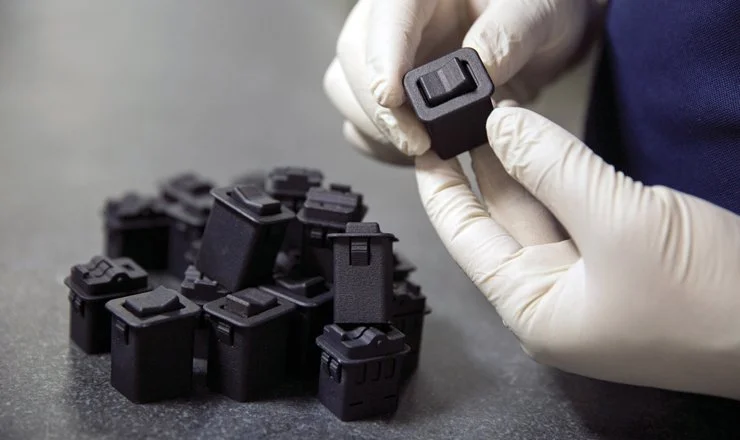 Gloved hands holding a small black 3D-printed polymer part among a group of similar components, illustrating additive manufacturing benefits.