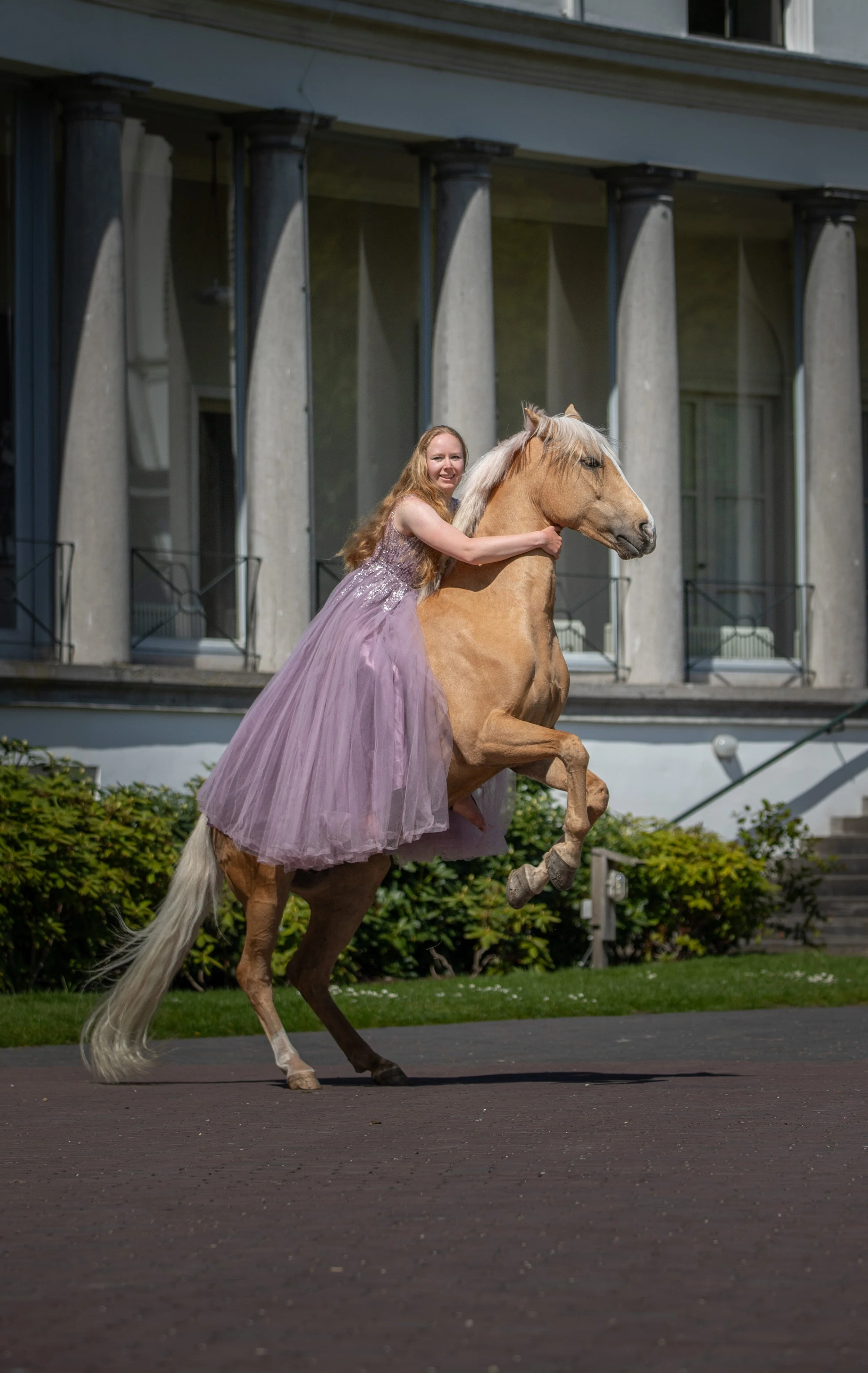 A woman in a purple dress riding a rearing horse in front of a large house with columns.