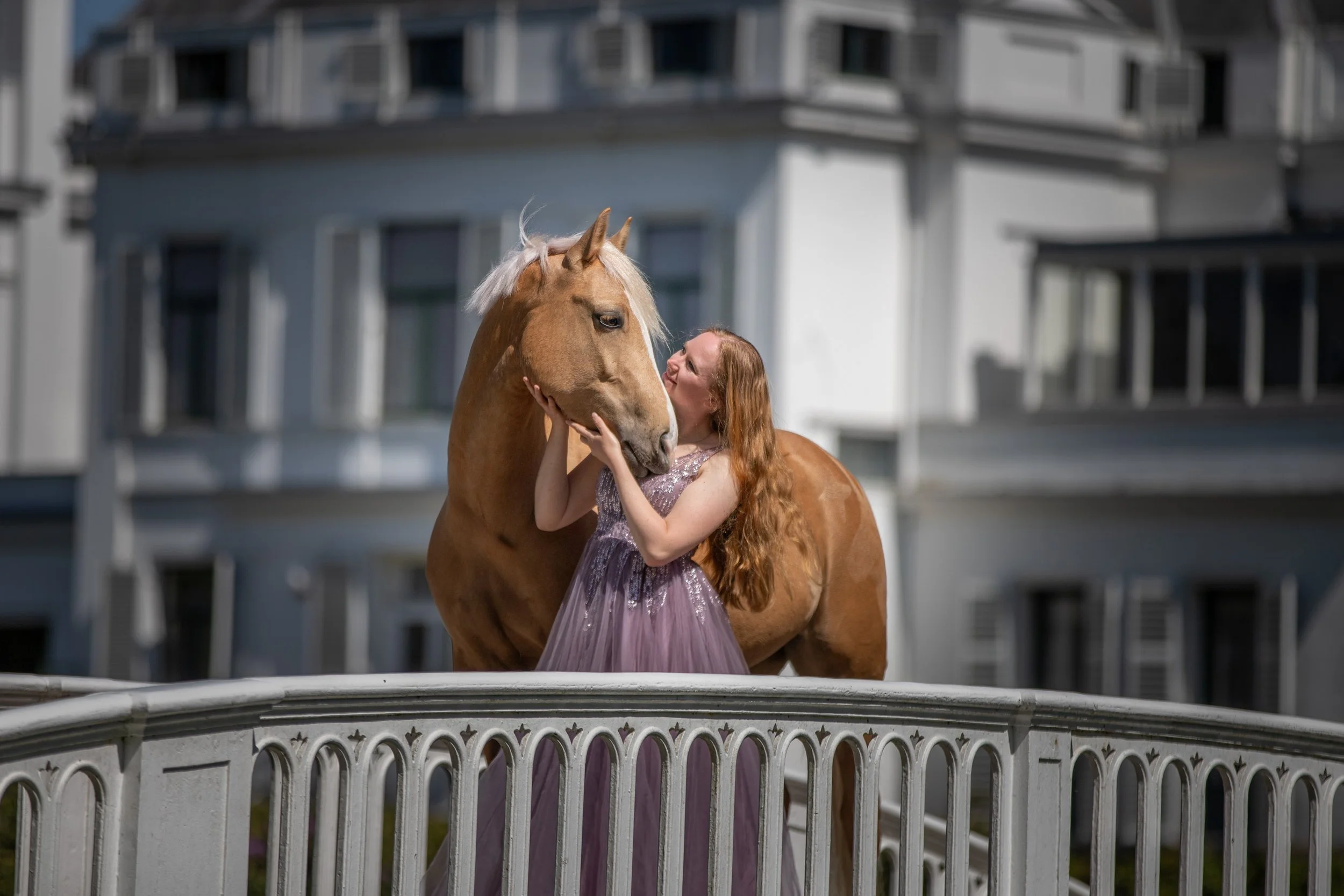 A woman with long red hair wearing a purple dress standing on a white ledge, kissing a large chestnut horse.