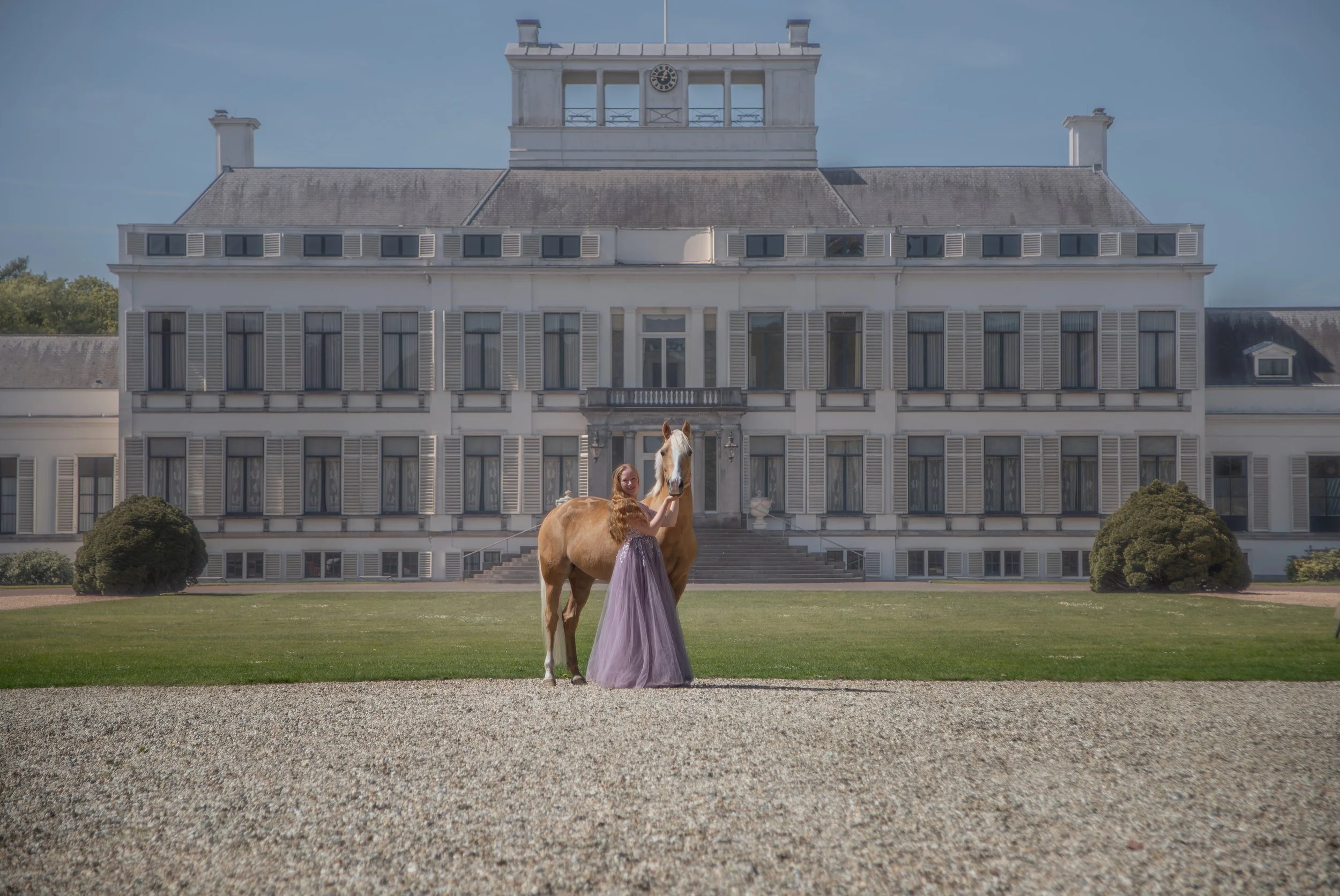 A woman in a long lavender gown standing next to a light brown horse with a white face in front of a large white mansion with multiple windows and a clock tower on top.