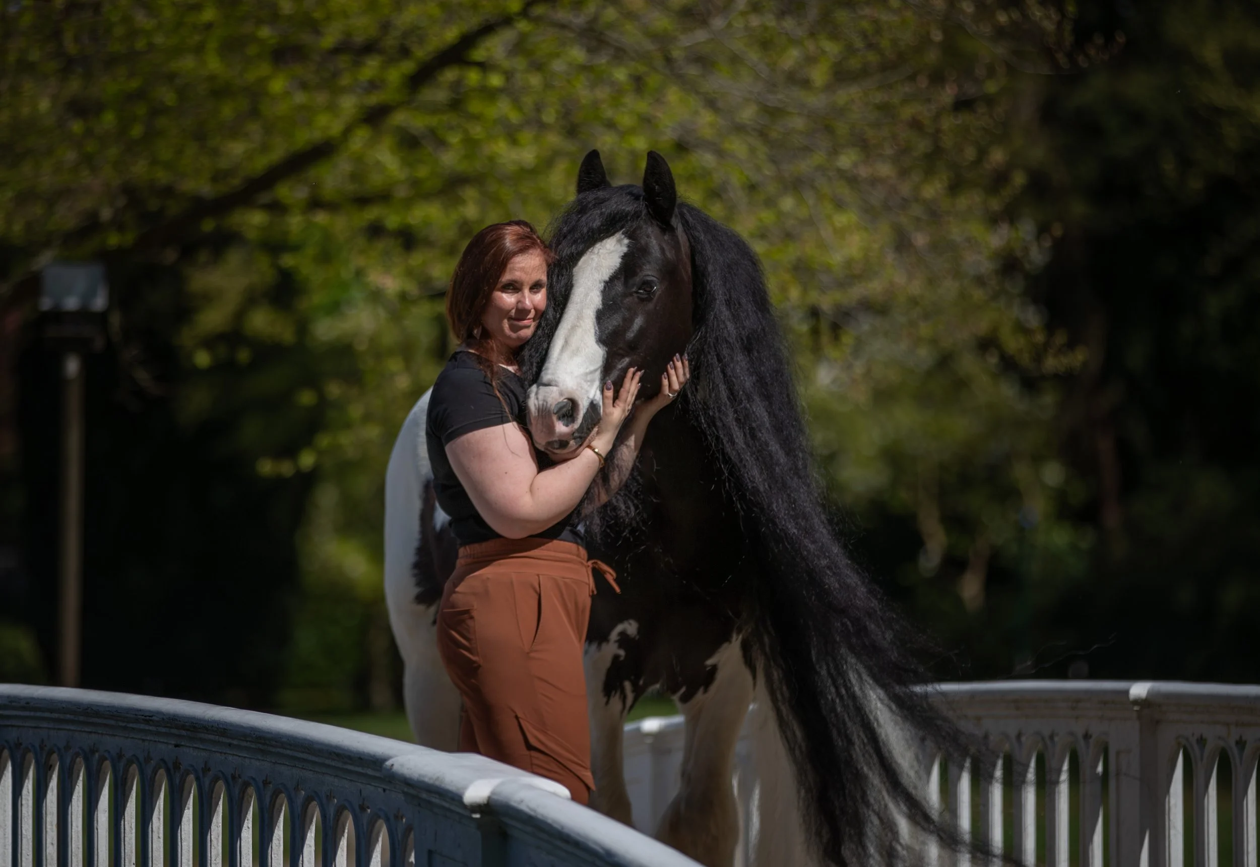 A woman with brown hair and a black shirt hugging a large black and white horse on a white bridge in a park.