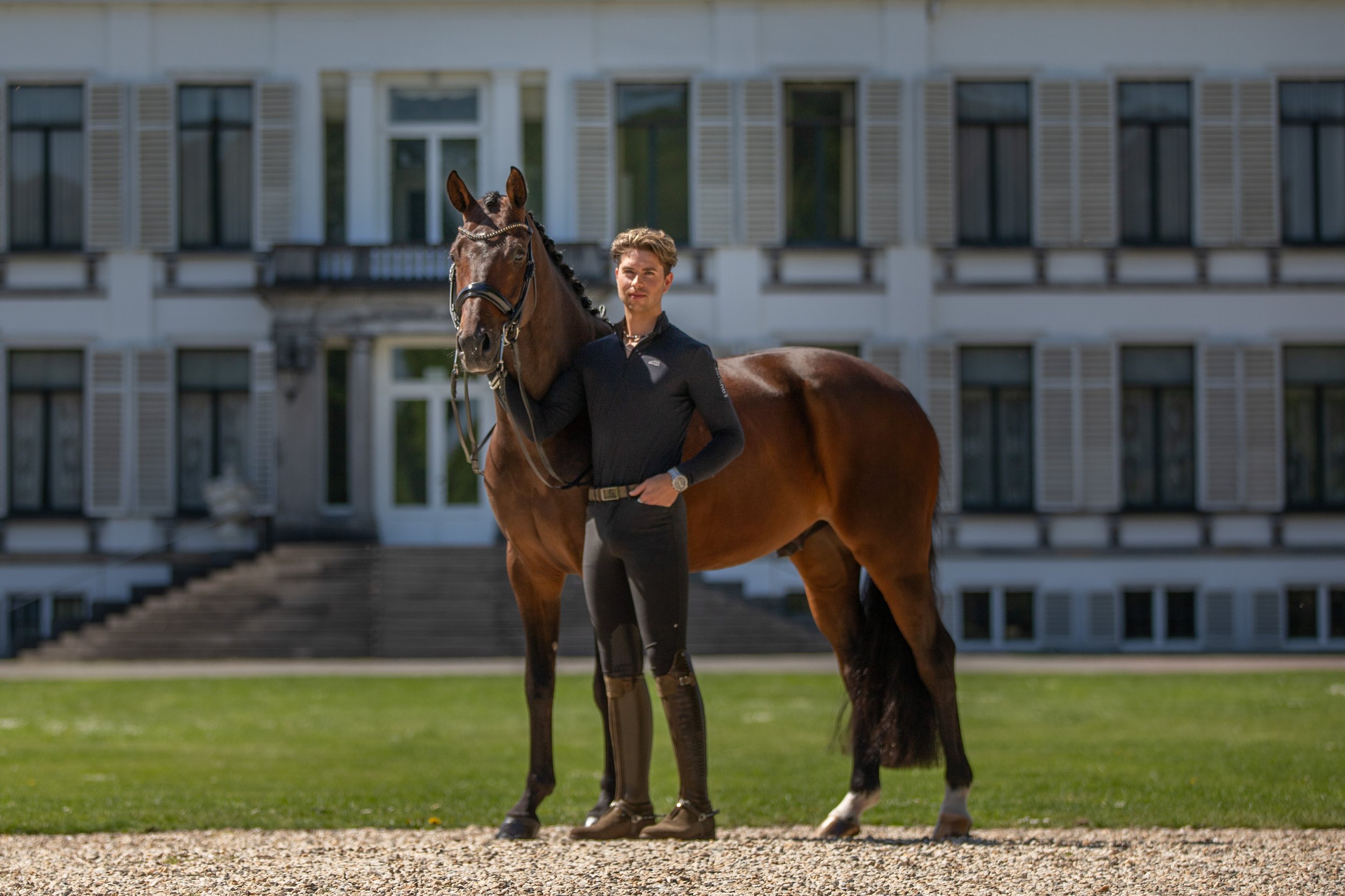 A man standing next to a brown horse on a gravel surface in front of a large white building with multiple windows and shutters.