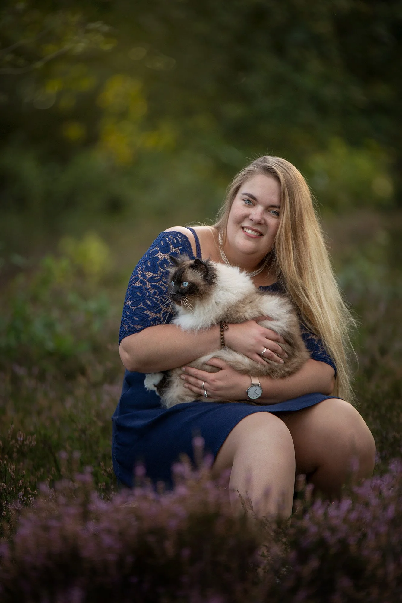 A woman sitting outdoors in purple flowers, holding a Siamese cat with blue eyes and a fluffy white and brown coat.