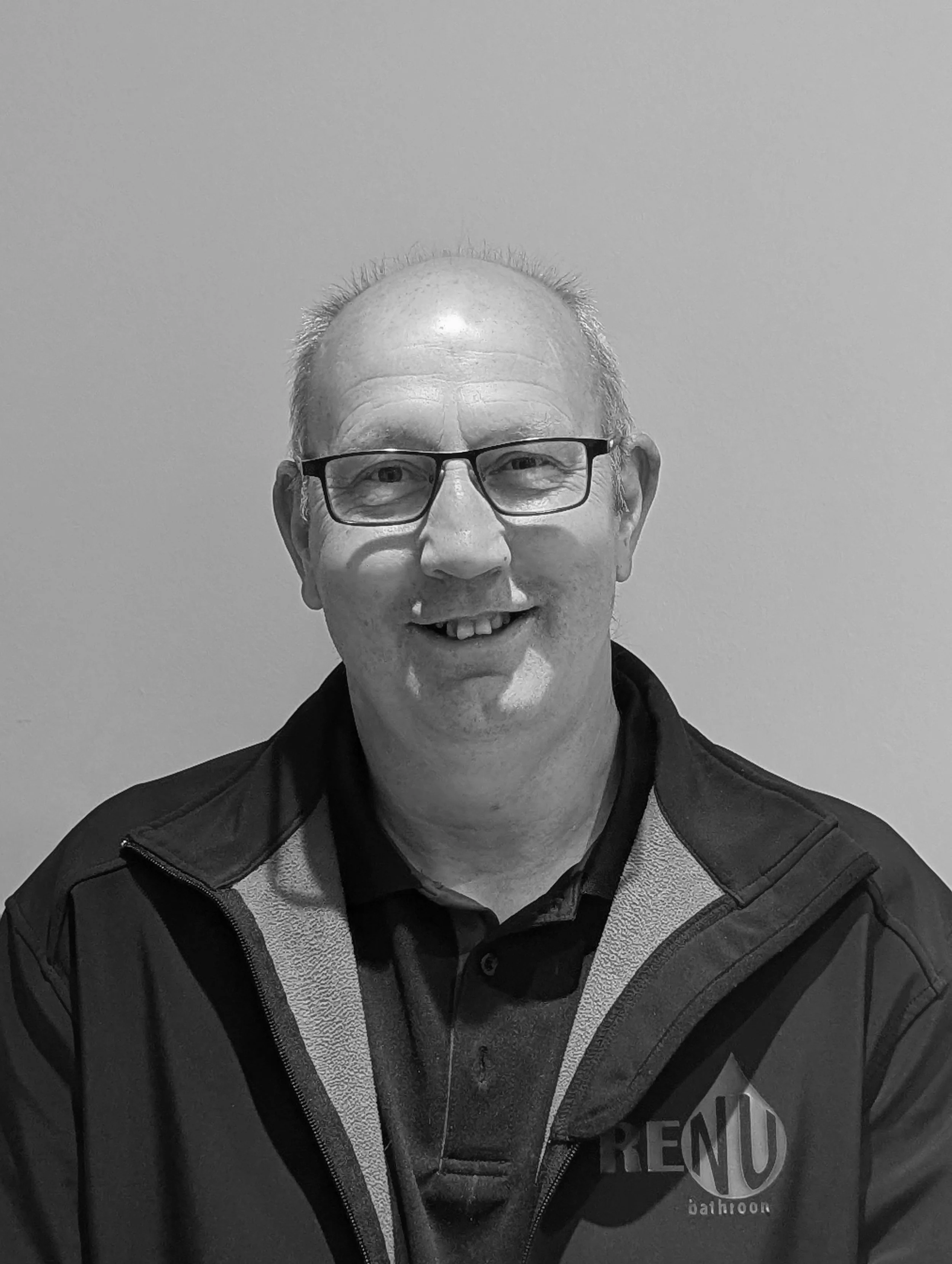 A smiling man with glasses in a black jacket and polo shirt, sitting against a plain background.