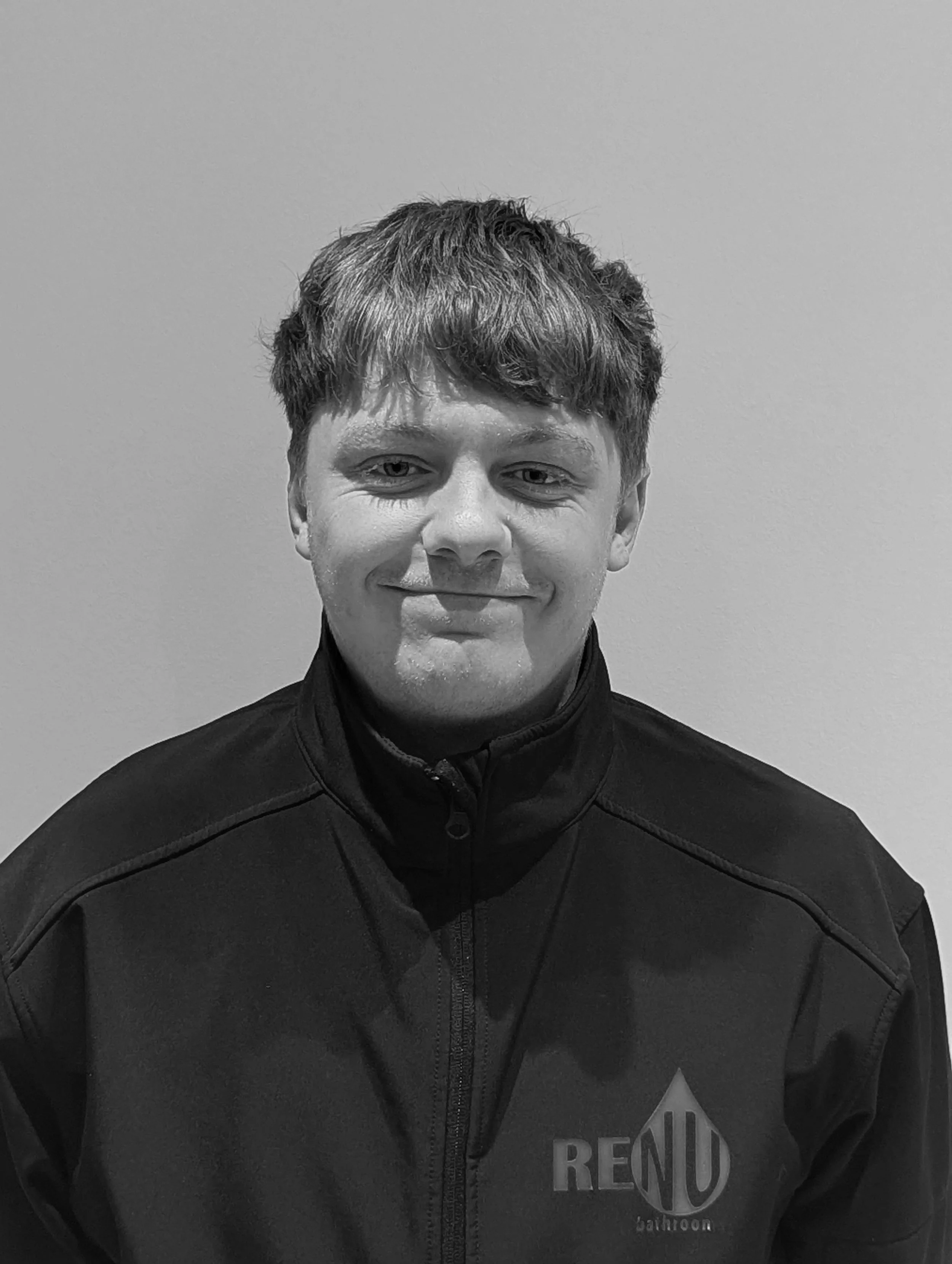 A young man with short hair wearing a black jacket with a logo that says 'RENU' on it, smiling softly, standing against a plain wall.