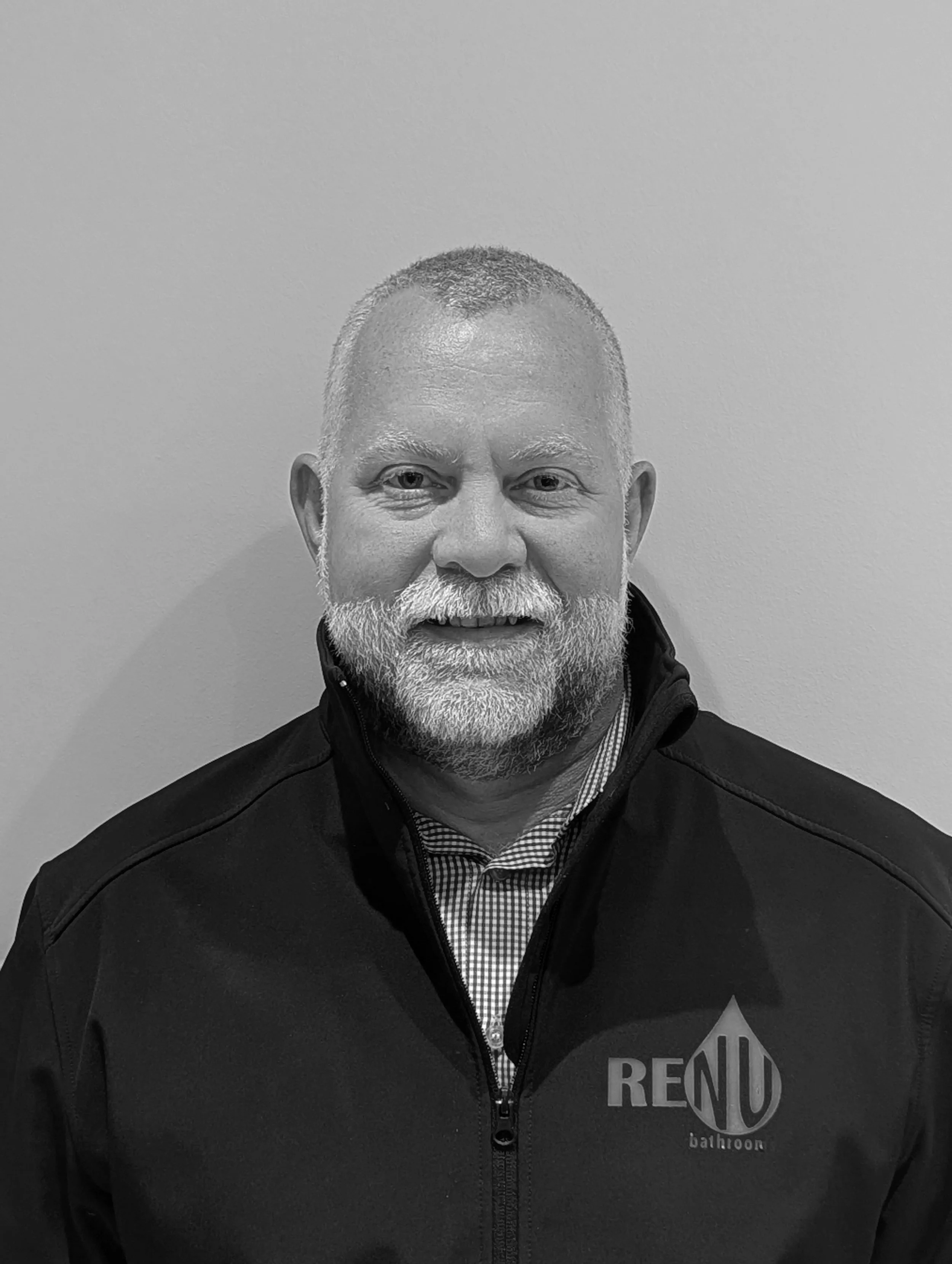 Black and white photo of a middle-aged man with short hair and a beard, wearing a black jacket with a 'RENU' logo and a checkered shirt underneath, standing against a plain wall.
